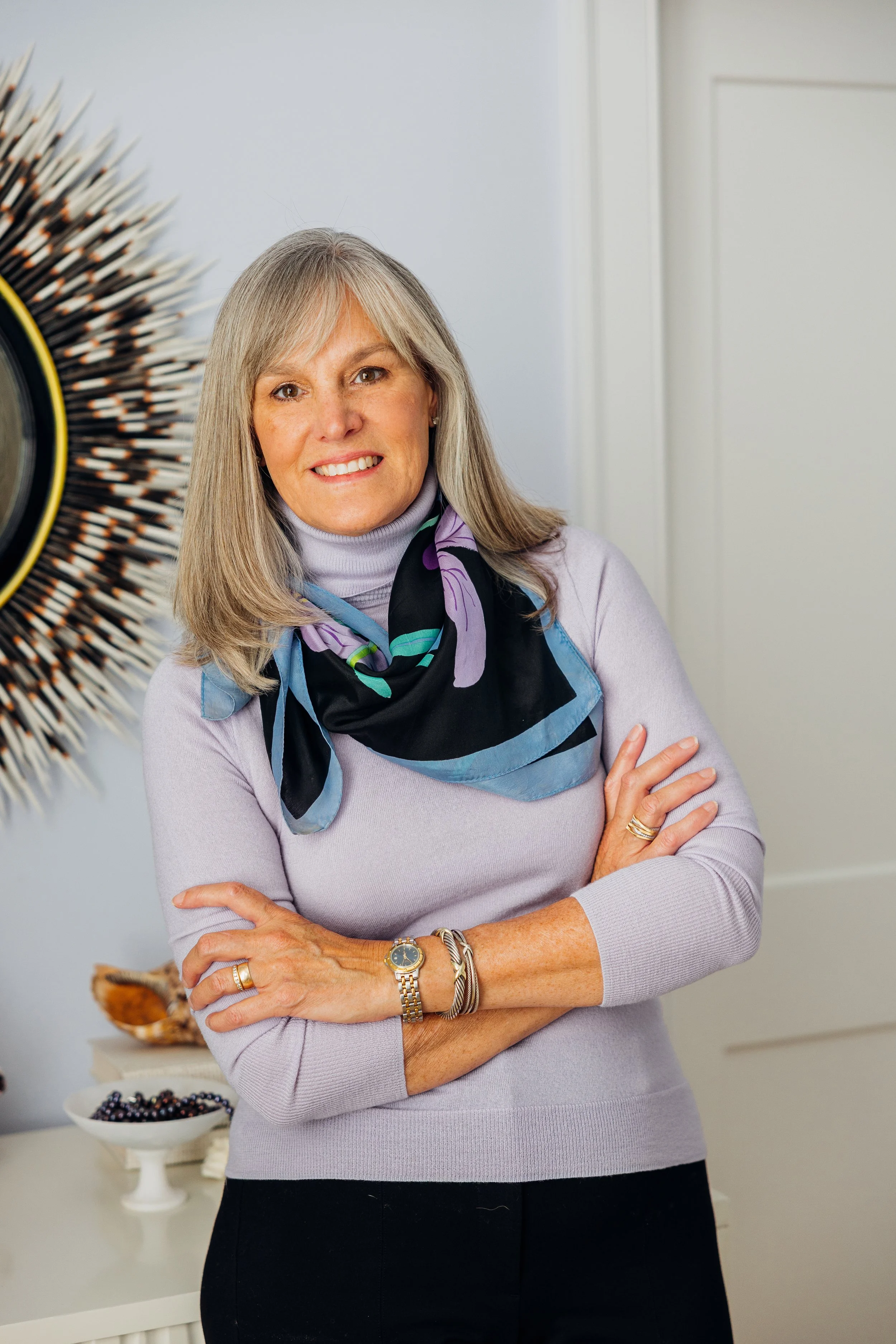 A woman with gray hair and a warm smile, wearing a lavender sweater and a black, purple, and teal scarf, standing with crossed arms in a decoratively furnished room.