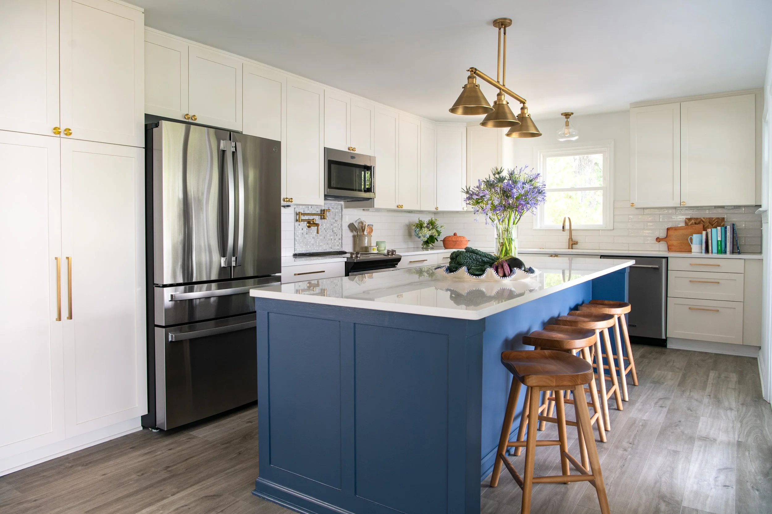 A modern kitchen with white cabinets, stainless steel refrigerator, black microwave, white tile backsplash, a blue island with wooden stools, and a bouquet of purple flowers on the counter near a window.