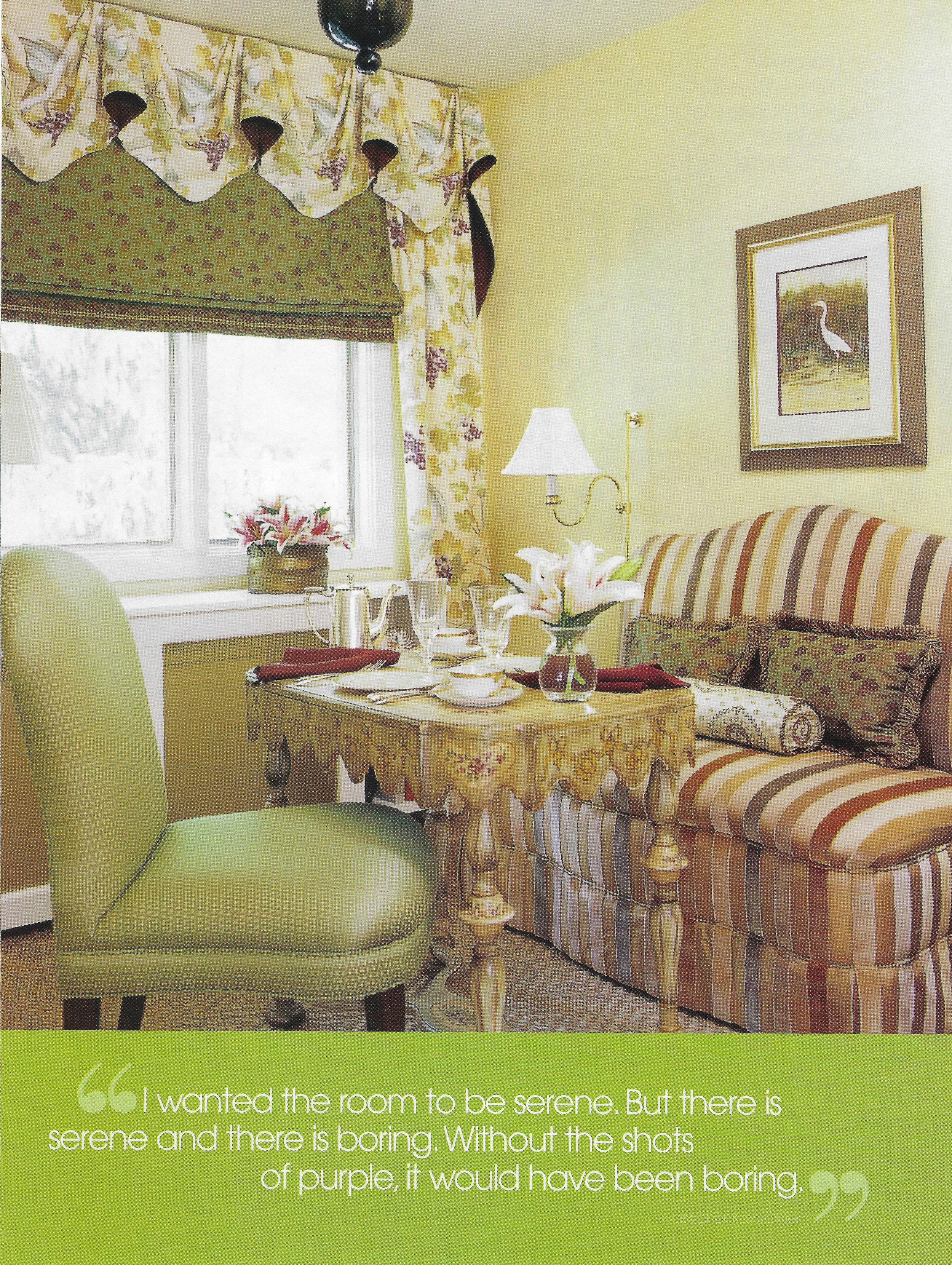 A cozy living room with a window covered by floral curtains, a yellow wall with a framed picture of a heron, a striped sofa with floral cushions, a yellow wooden table with a vase of lilies and tableware, a green upholstered chair, a lamp, and a floral arrangement on the windowsill.