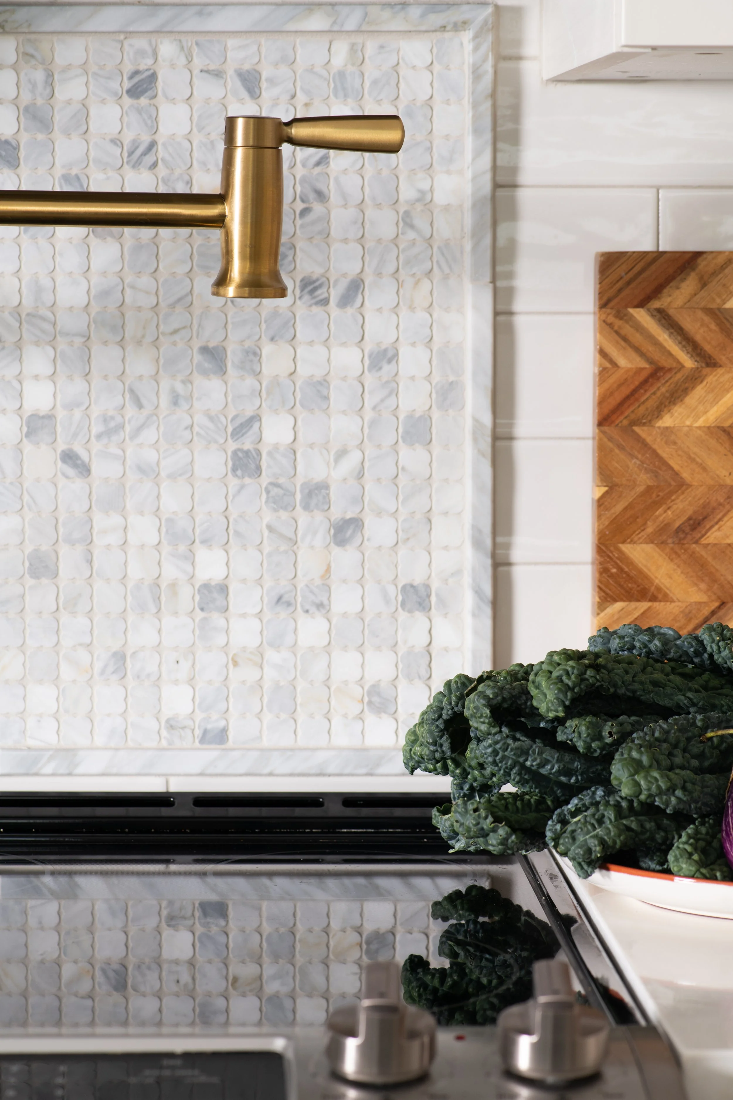 A kitchen backsplash made of small, clover shaped white and gray marble tiles, with brass pot filler and a chevron-patterned cutting board. The stove's black glass surface is reflected in the lower part of the image.