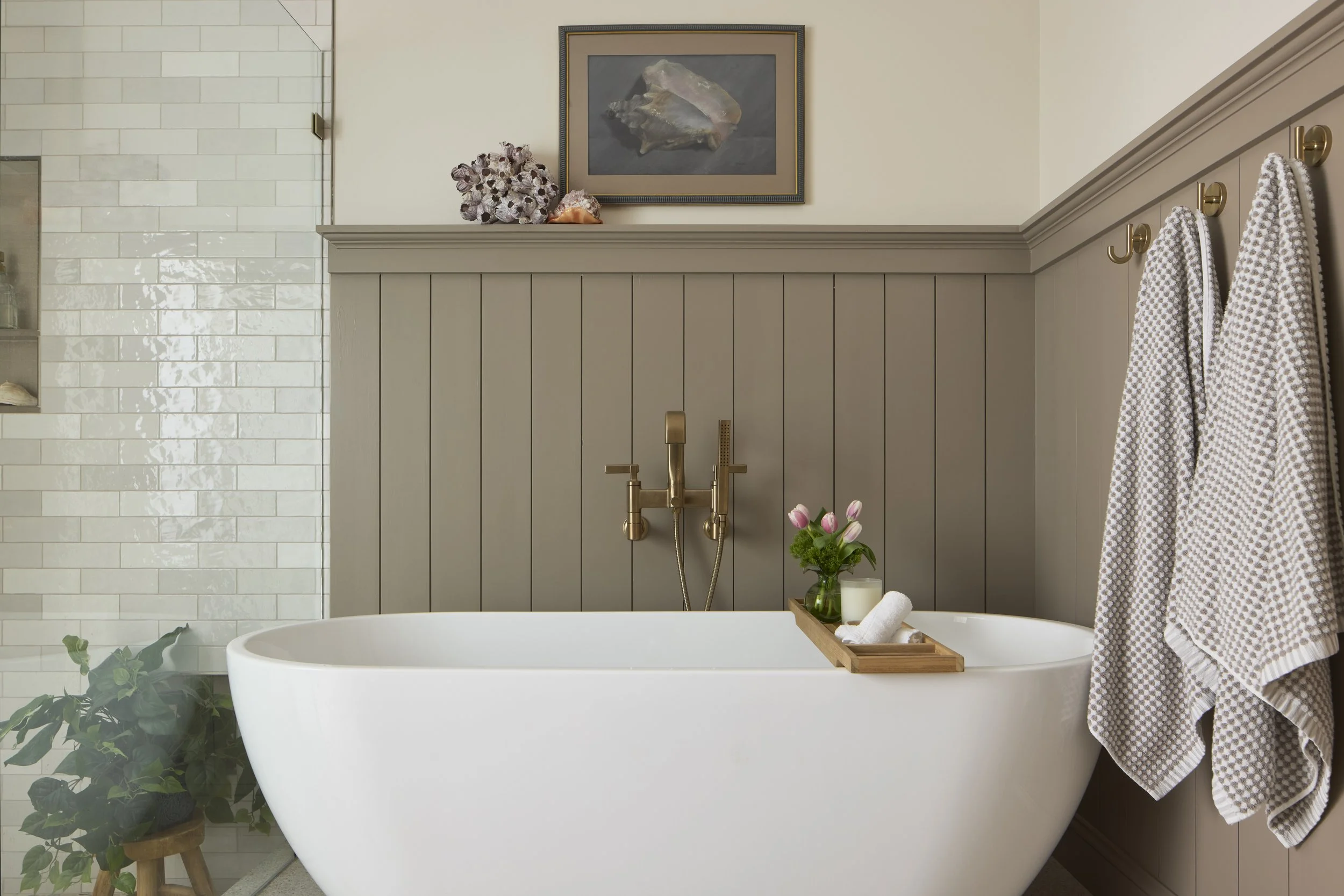 Modern bathroom with white freestanding bathtub, wall-mounted brass faucet, painted wainscoting, framed artwork, towels hanging on hooks, tub tray with flowers and candle, next to tiled walk in shower.