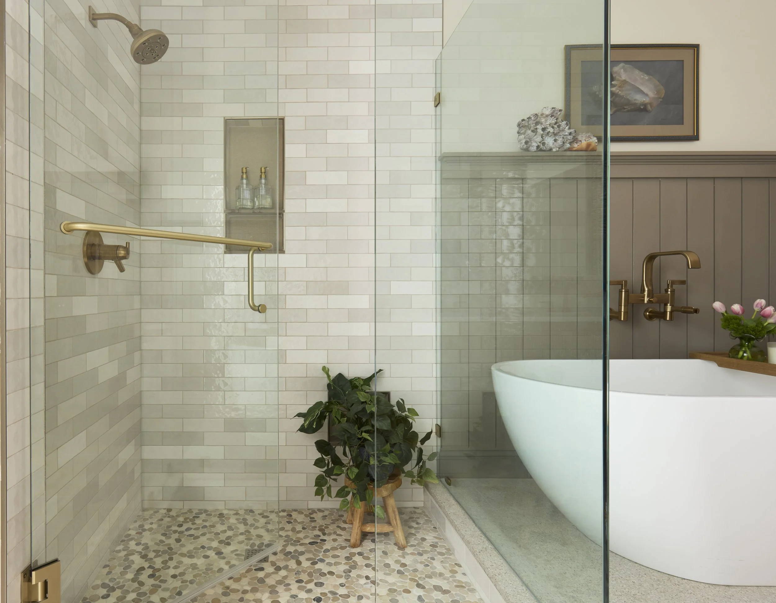Modern bathroom with a separate glass-enclosed shower area on the left featuring cream-colored subway tiles, a showerhead, brass fixtures, a small niche with bottles, and a pebble tile floor. On the right, a freestanding white bathtub with brass fixtures, with a wooden ledge.