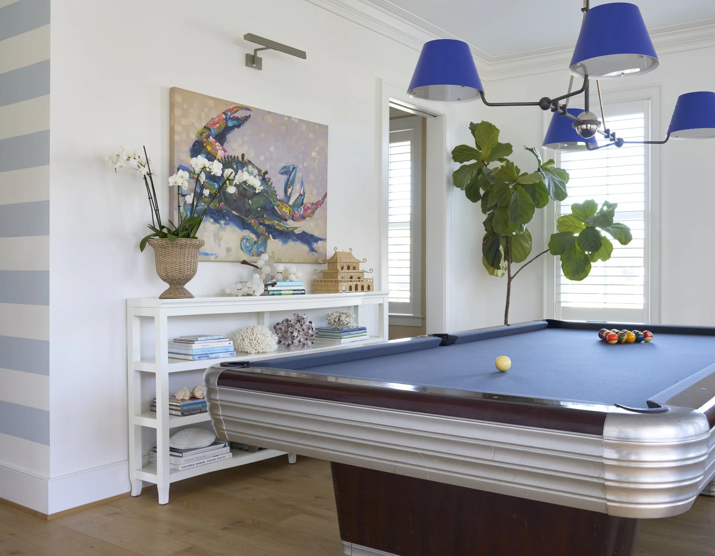 A room with a pool table, a blue lampshade chandelier, a large indoor plant near windows with shutters, a white bookshelf with decorative items, a colorful artwork of a crab above the bookshelf, and a potted orchid on top of the bookshelf.