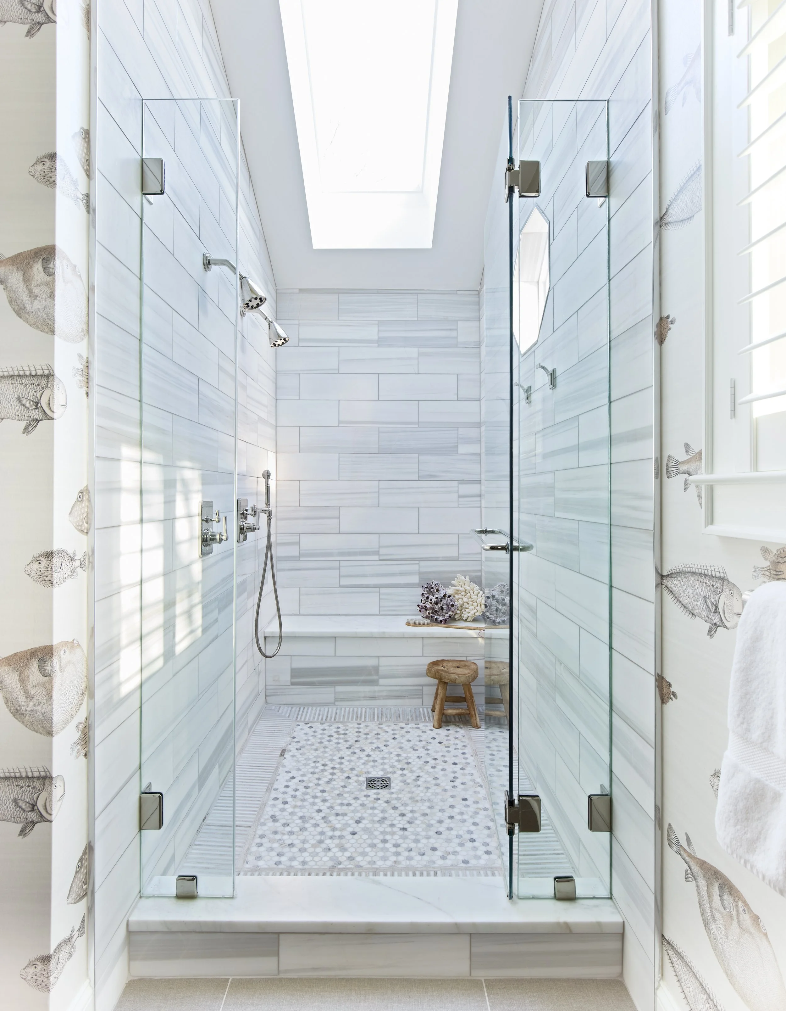 View of a bright, modern shower with glass doors, marble tile walls, a skylight, and a bench with coral decorations and a small wooden stool.