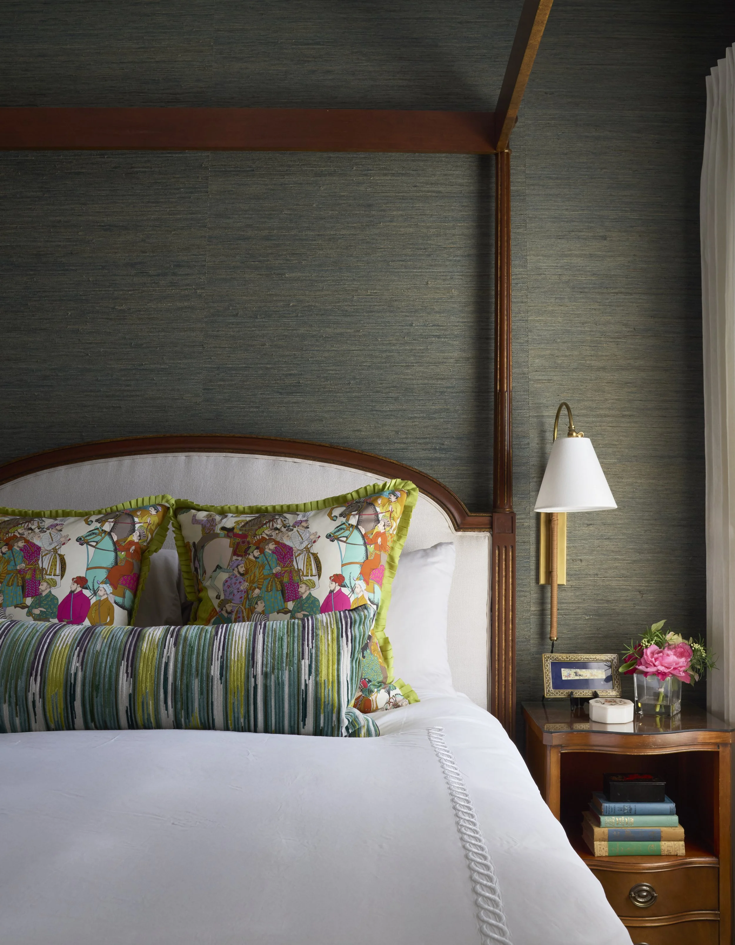 A neatly made bed with decorative pillows featuring colorful, artistic scenes. A bedside table with books, a framed picture, a flower vase with pink flowers, and a wall-mounted lamp are visible, set against a textured greenish wall in a cozy room.