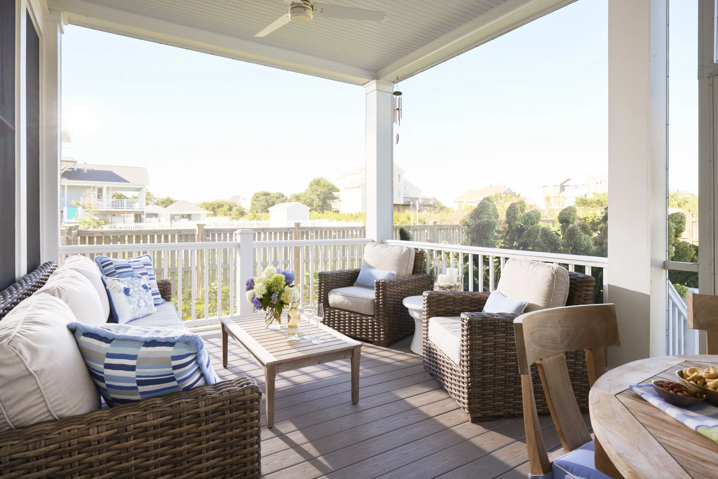 Covered porch with wicker and cushioned seating, a coffee table with flowers and drinks, overlooking a backyard with trees and neighboring houses