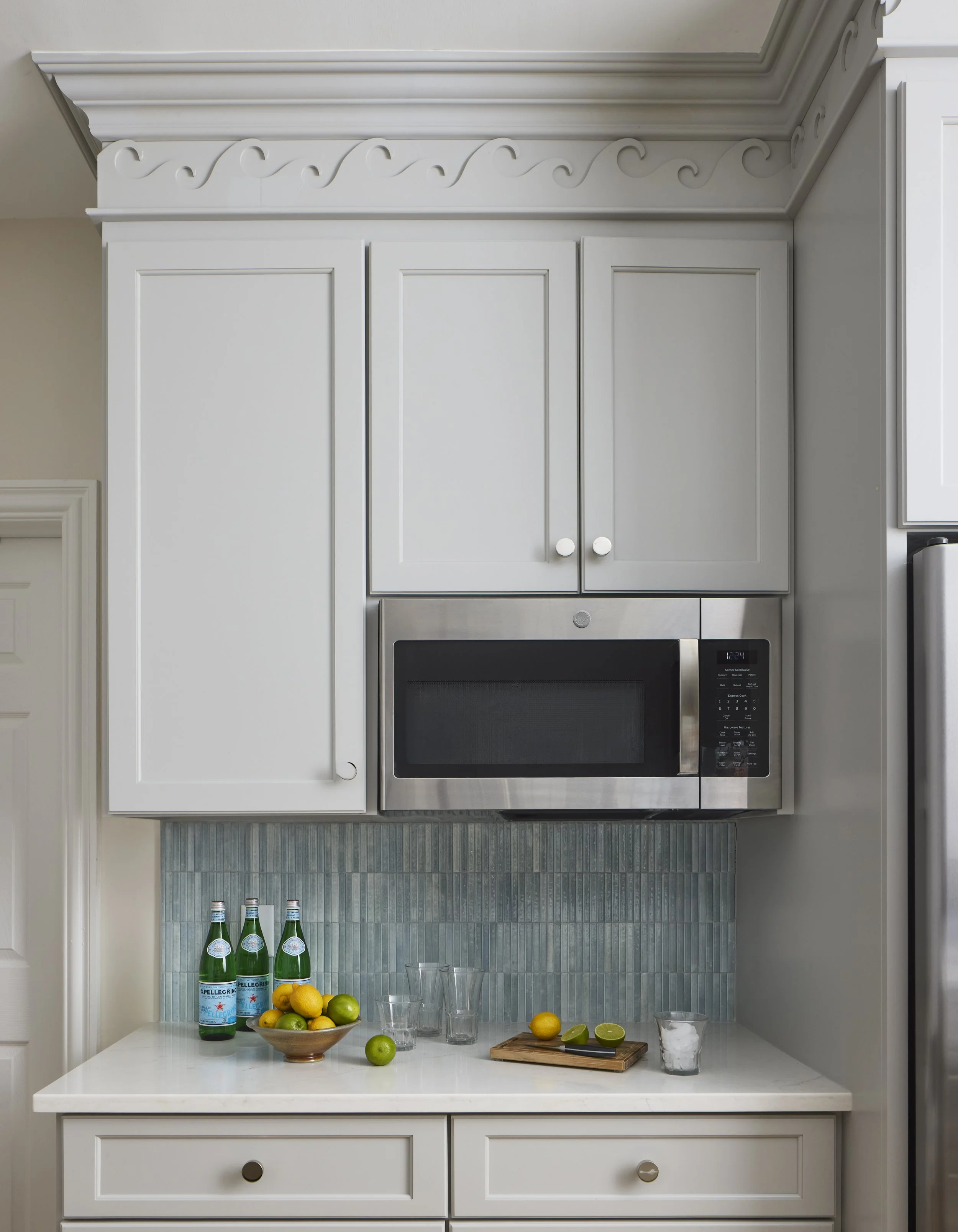 Gray kitchen cabinets with a built-in microwave, a countertop with bottles of sparkling water, a bowl of lemons and limes, some glasses, a cutting board with a lemon and lime, and a glass of ice.