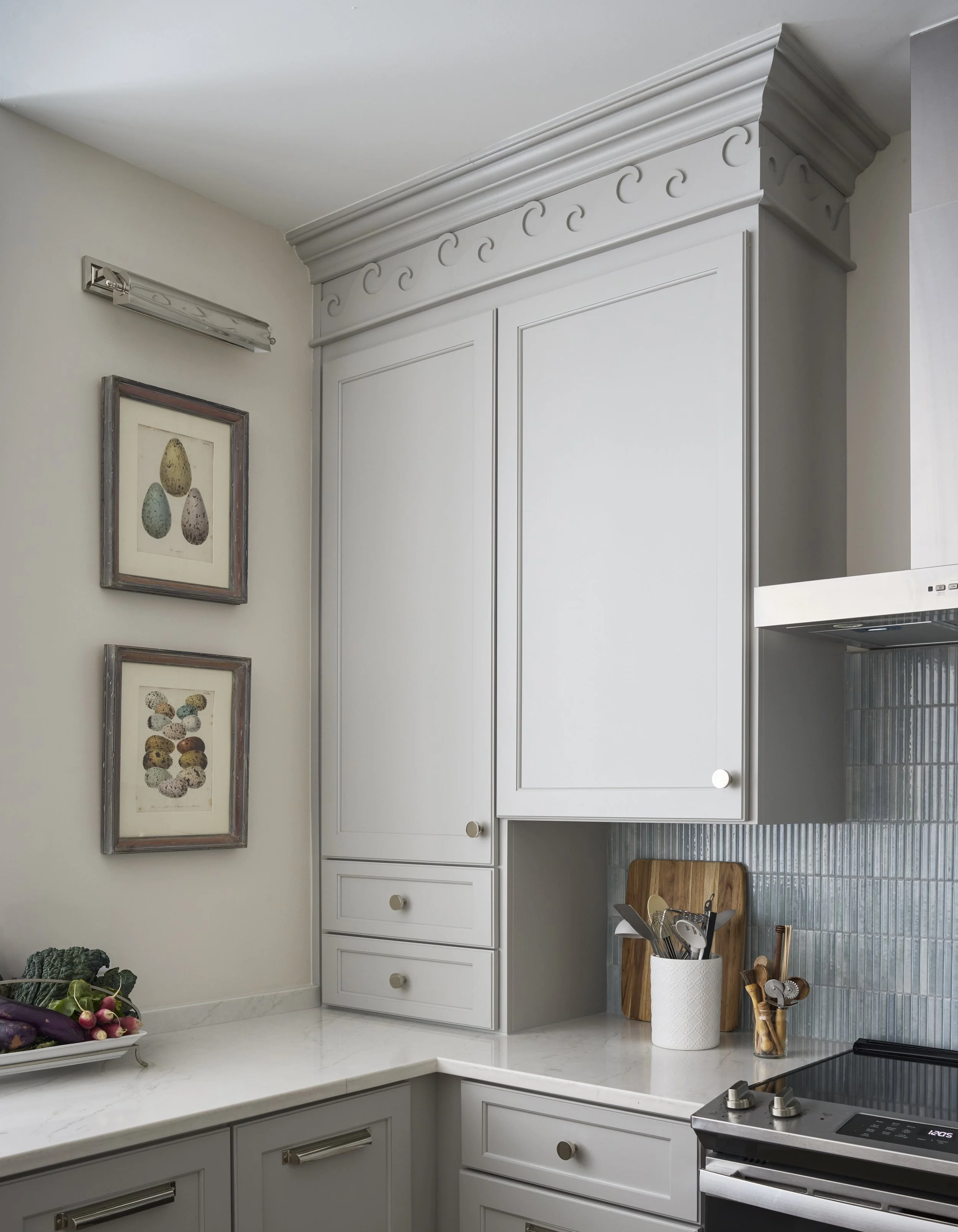 A section of a kitchen with gray cabinets, a marble countertop, framed artwork of eggs and other items, and kitchen utensils in a white container, with a cutting board and a stovetop.