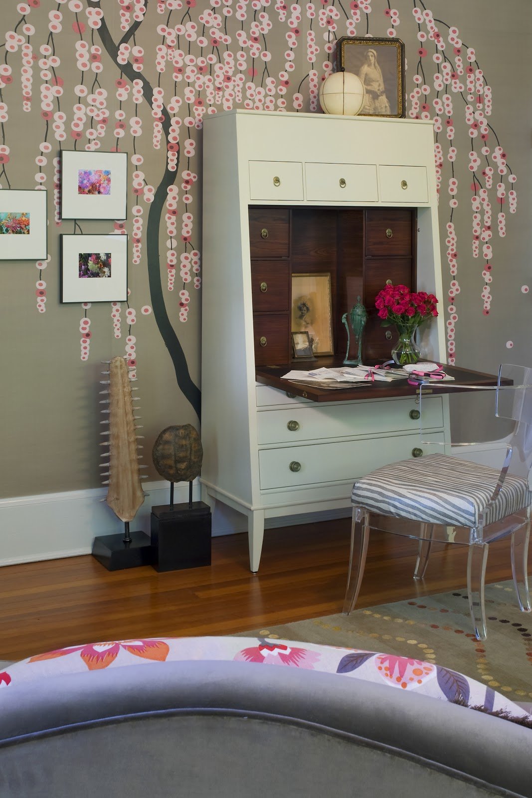 Decorative interior featuring a floral mural wall with pink blossoms, a white and wood secretary with decorative items and books, a chair with a striped cushion, a vase of pink flowers, and framed artwork.