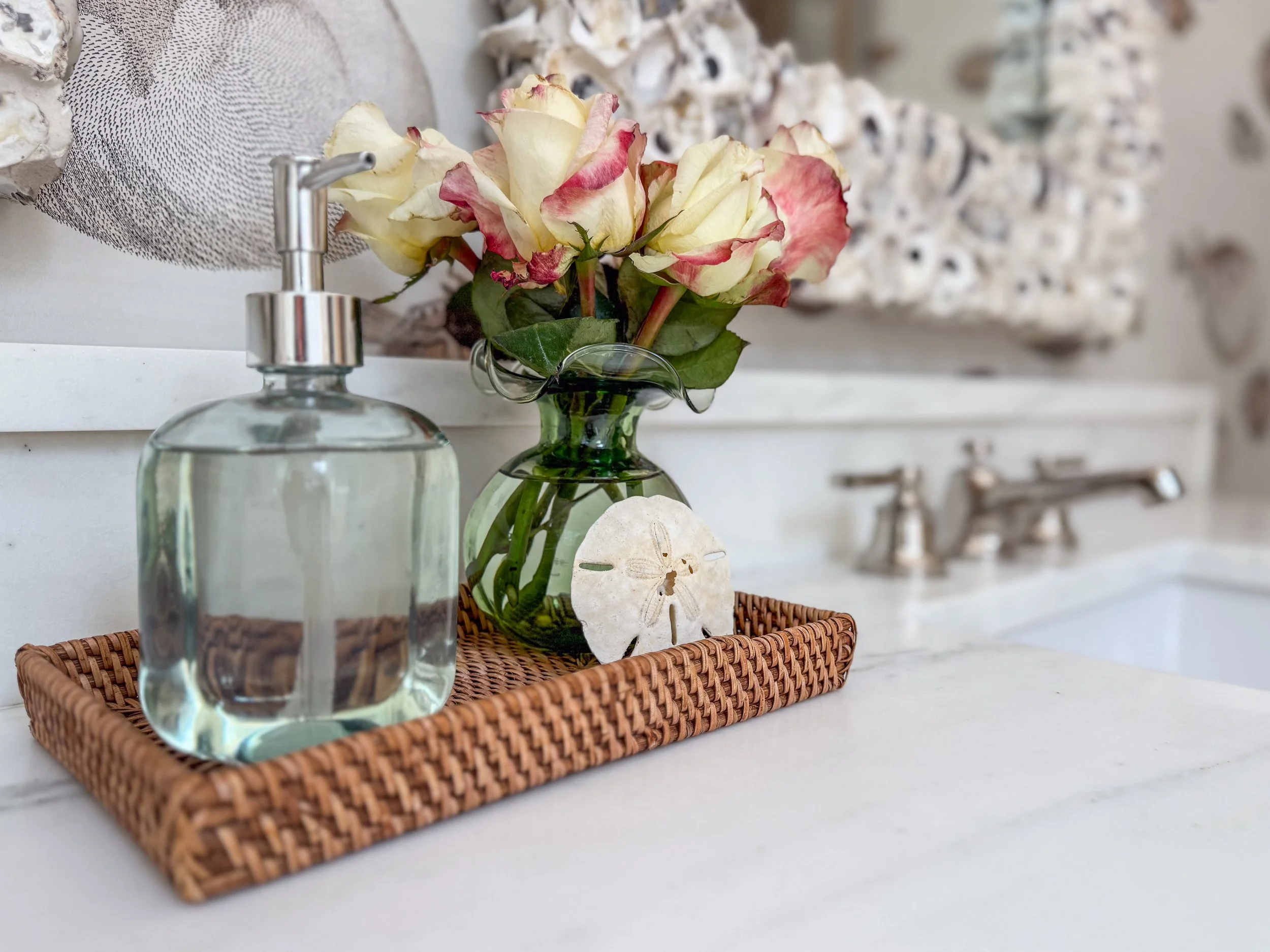 A clear soap dispenser, a glass vase with pink and cream roses, and a piece of dried plant on a woven tray on a white countertop.