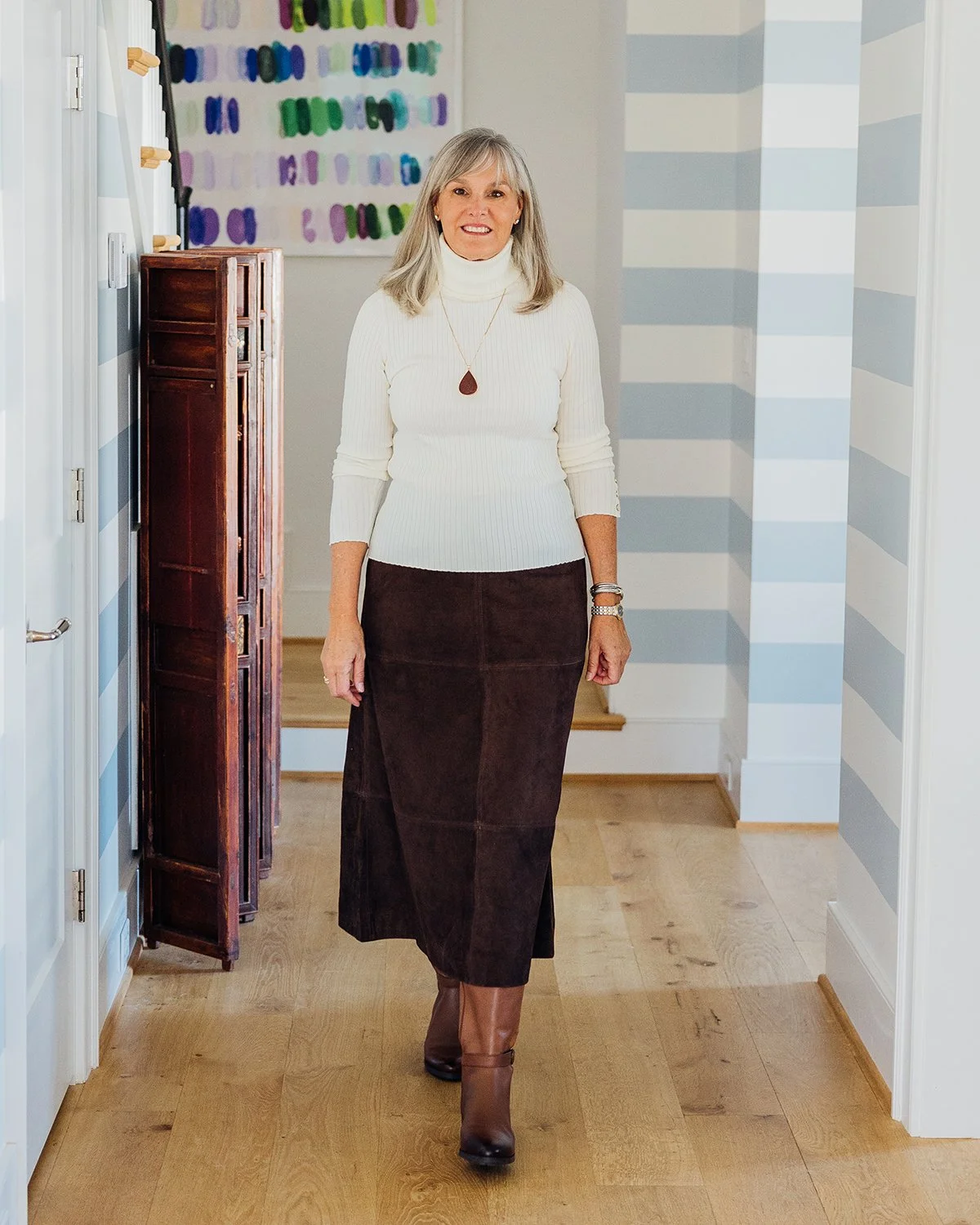 A woman with gray hair and a smile, wearing a white turtleneck sweater, a long dark brown skirt, and brown ankle boots, standing in a hallway with wooden flooring and striped blue and white walls.