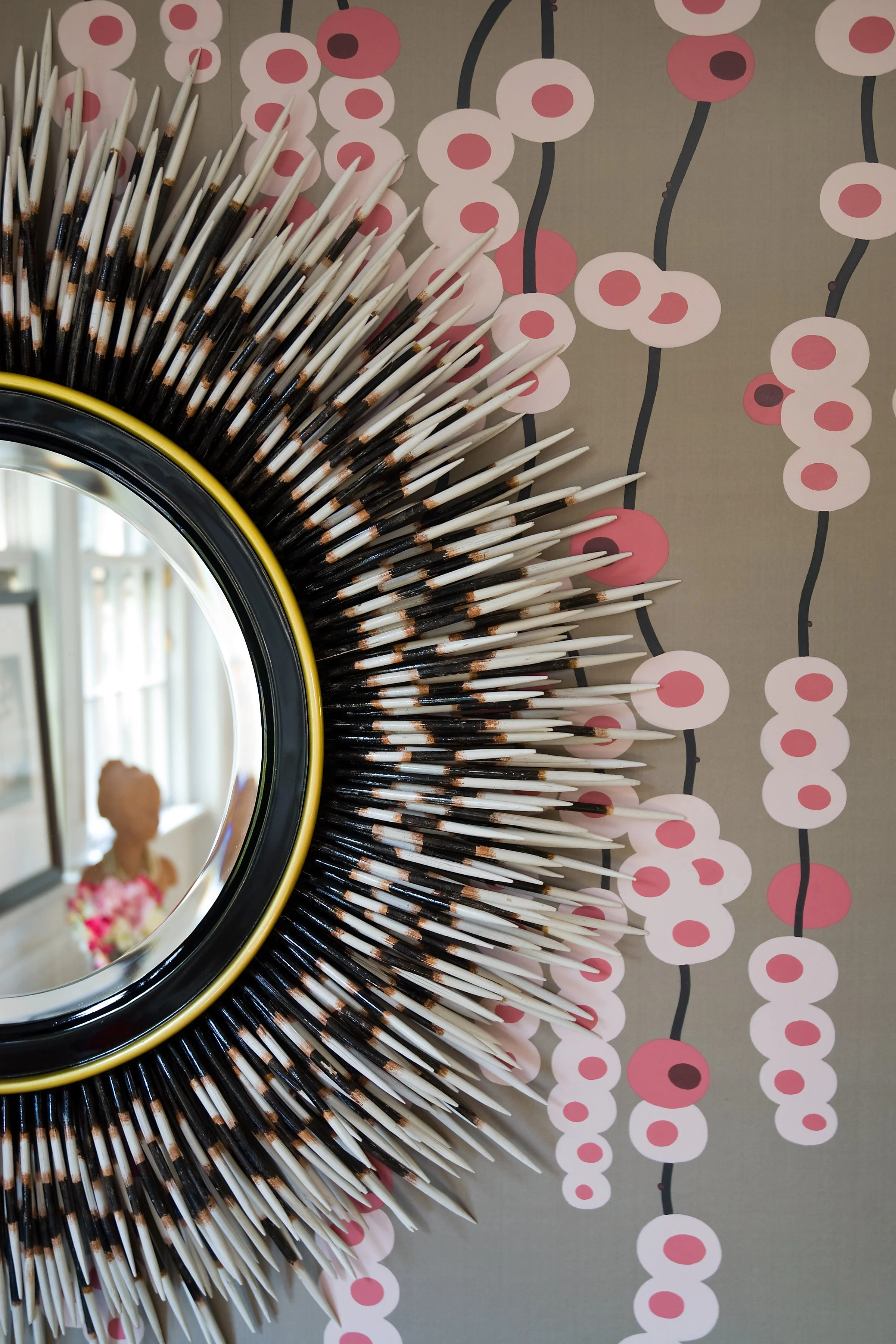A decorative mirror with a frame made of porcupine quills, mounted on a wall with pink and white circular patterned wallpaper.