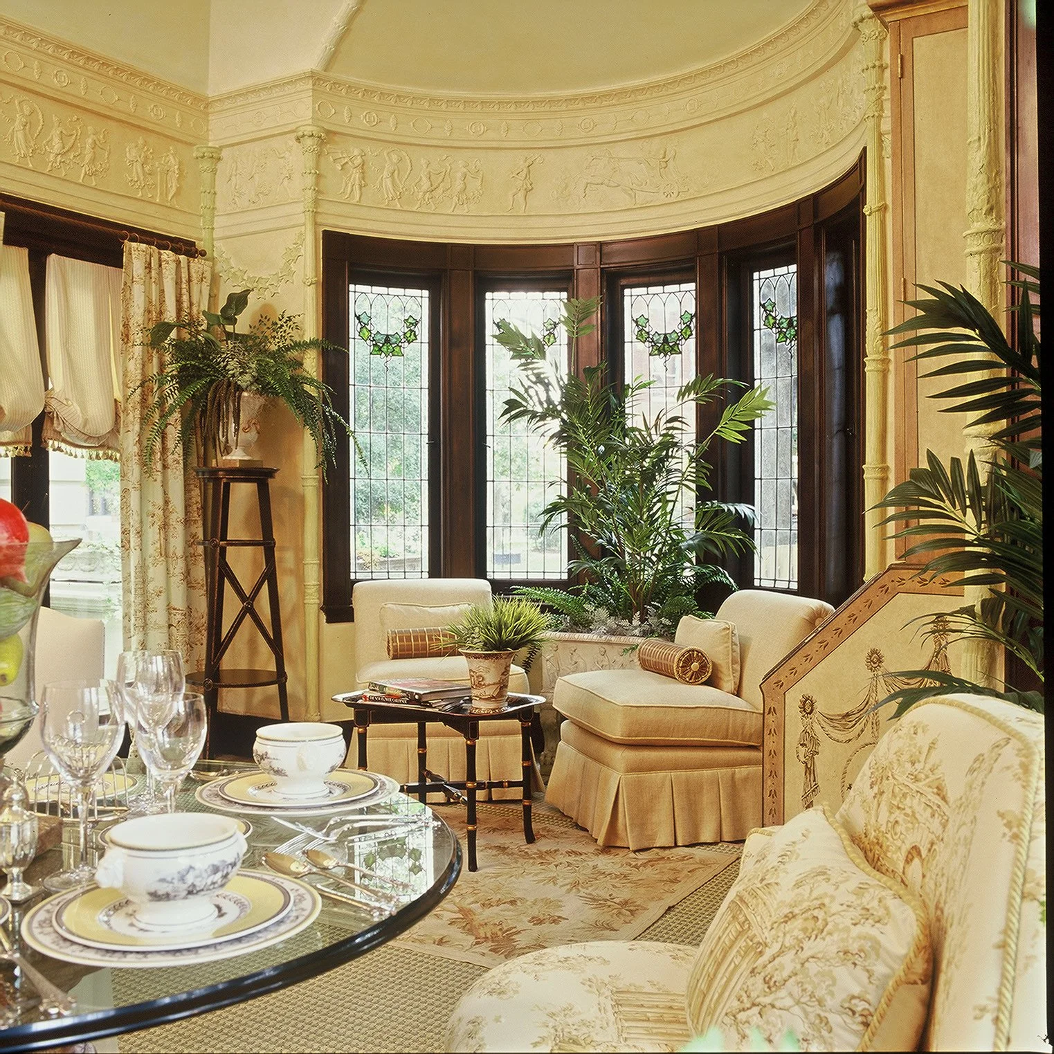 Elegant living room with large bay window, cream-colored tufted sofa, armchair, decorative pillows, and potted plants, including a tall fern and a palm, with vintage-style curtains.
