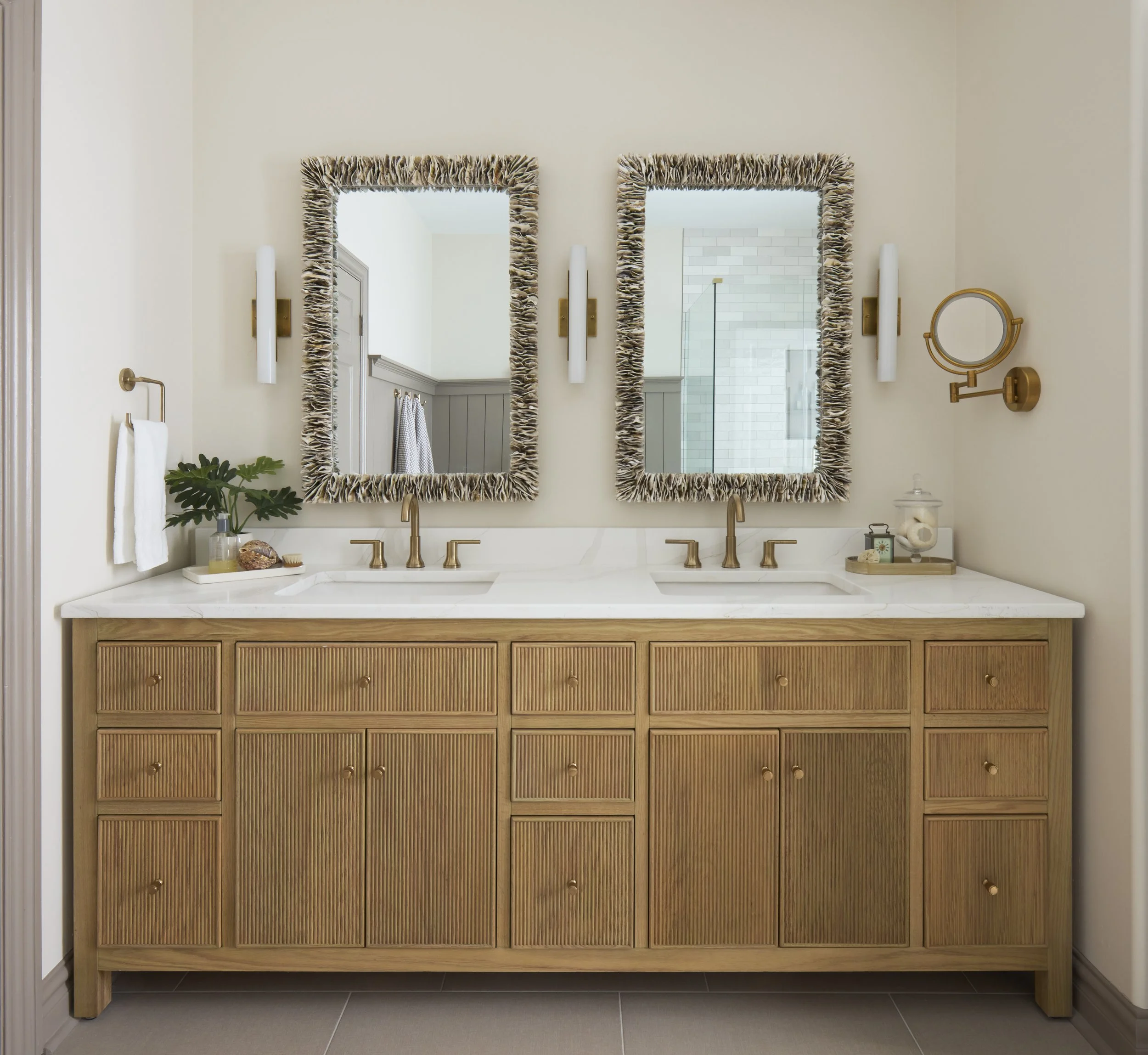 Bathroom vanity with two oyster shell mirrors, brass fixtures, marble countertop, fluted panel vanity, and decor items including a plant, soap dispenser, brass make up mirror, and decorative shell accessories.