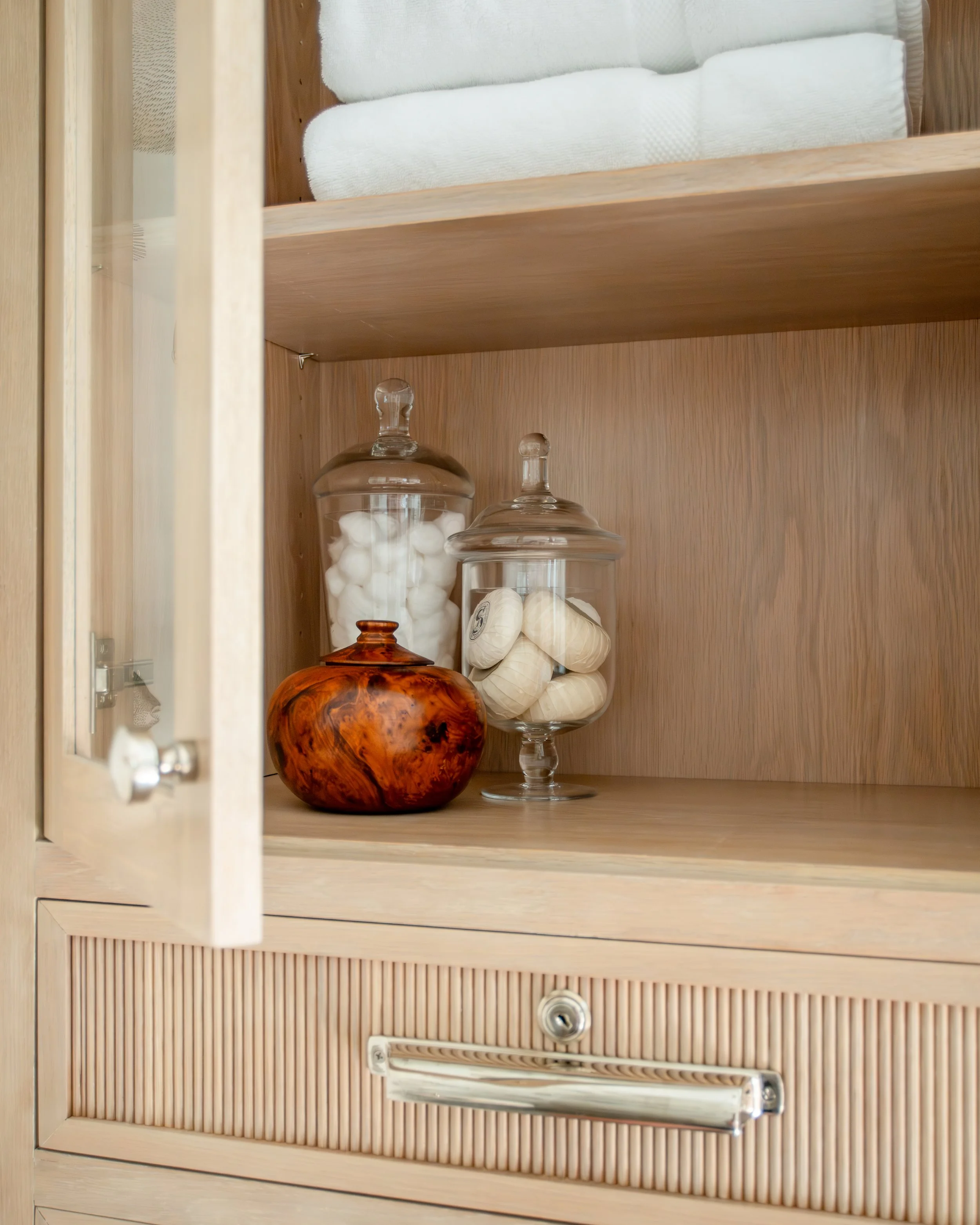 Inside a wooden cabinet, two glass jars filled with white cotton balls and cotton rounds are on display, with a decorative orange-brown wooden container next to them.