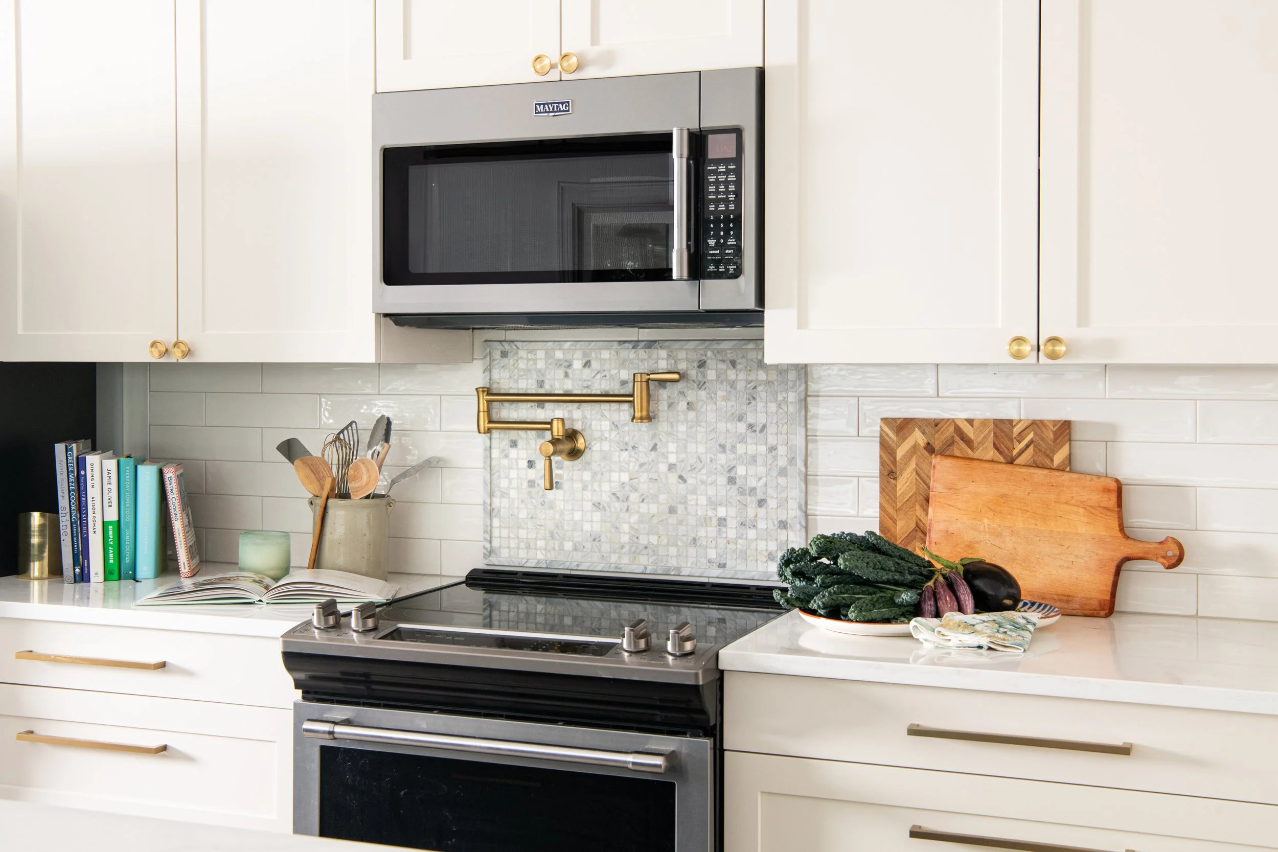 Kitchen with white cabinets, a black stove, stainless steel microwave, gold pot filler faucet, mosaic tile backsplash, cutting board, vegetables, and books on the countertop.