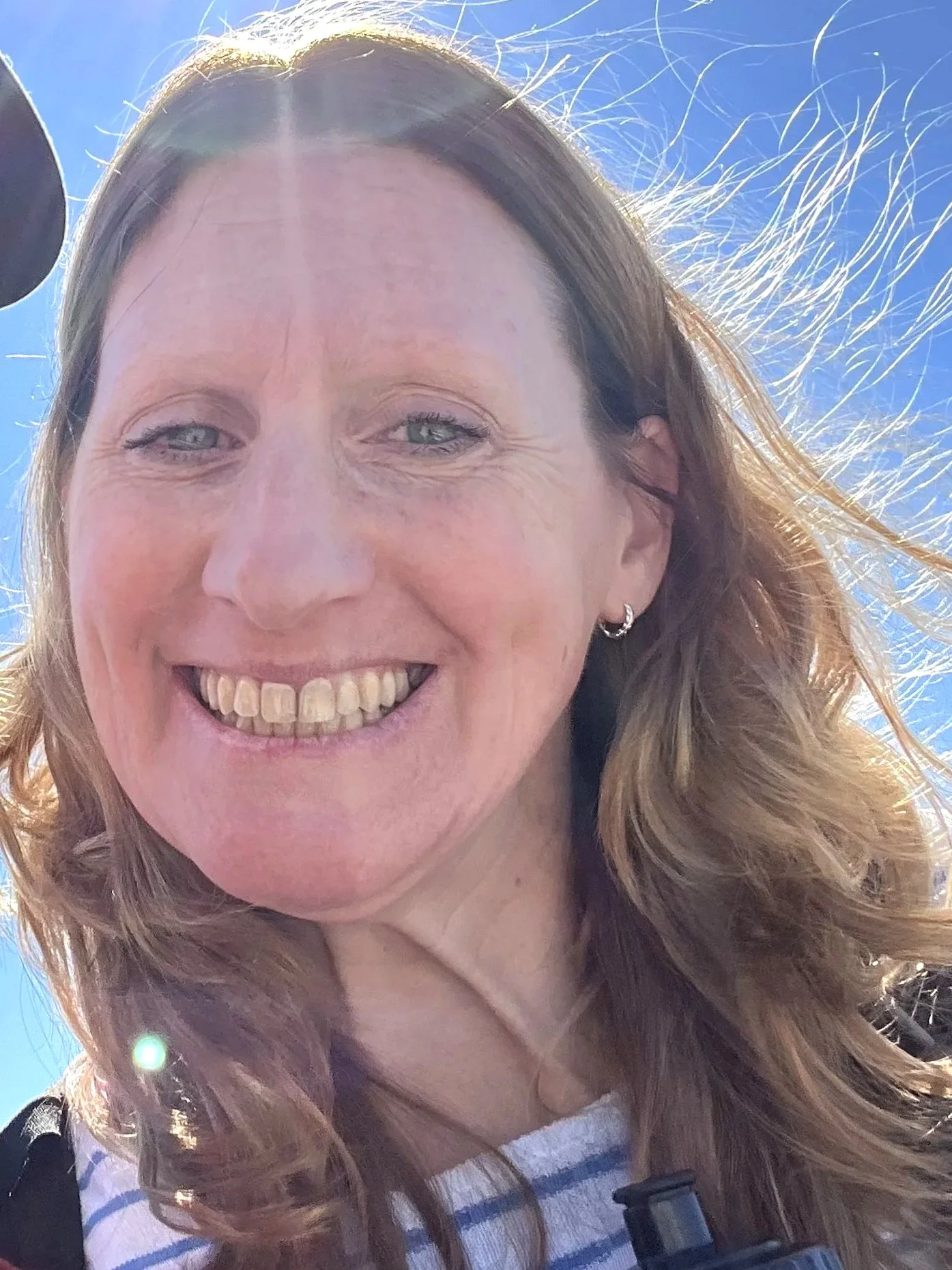 A woman with wavy light brown hair smiling outdoors against a blue sky.
