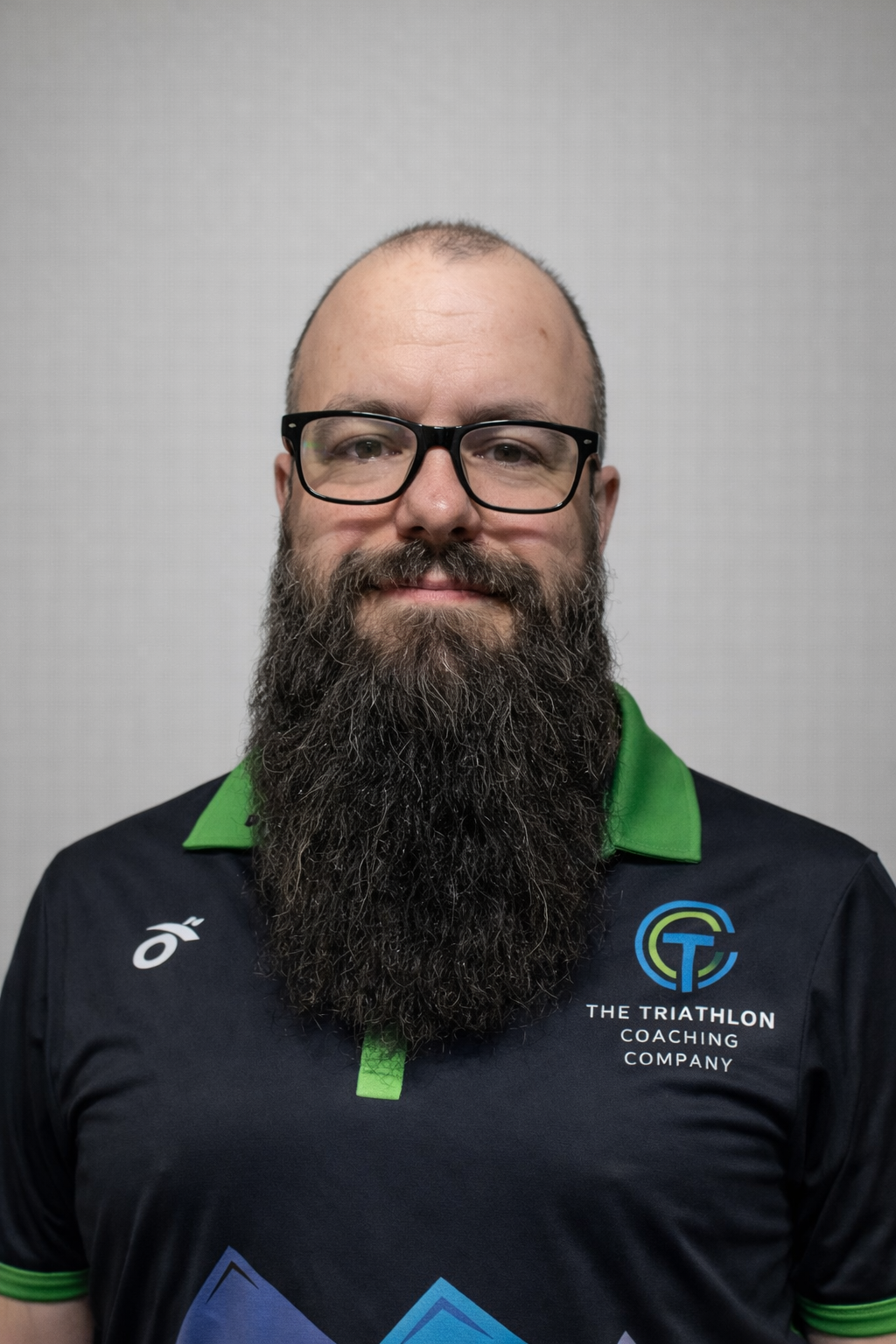 A man with glasses and a large beard wearing a black and green sports shirt with logo for The Triathlon Coaching Company.