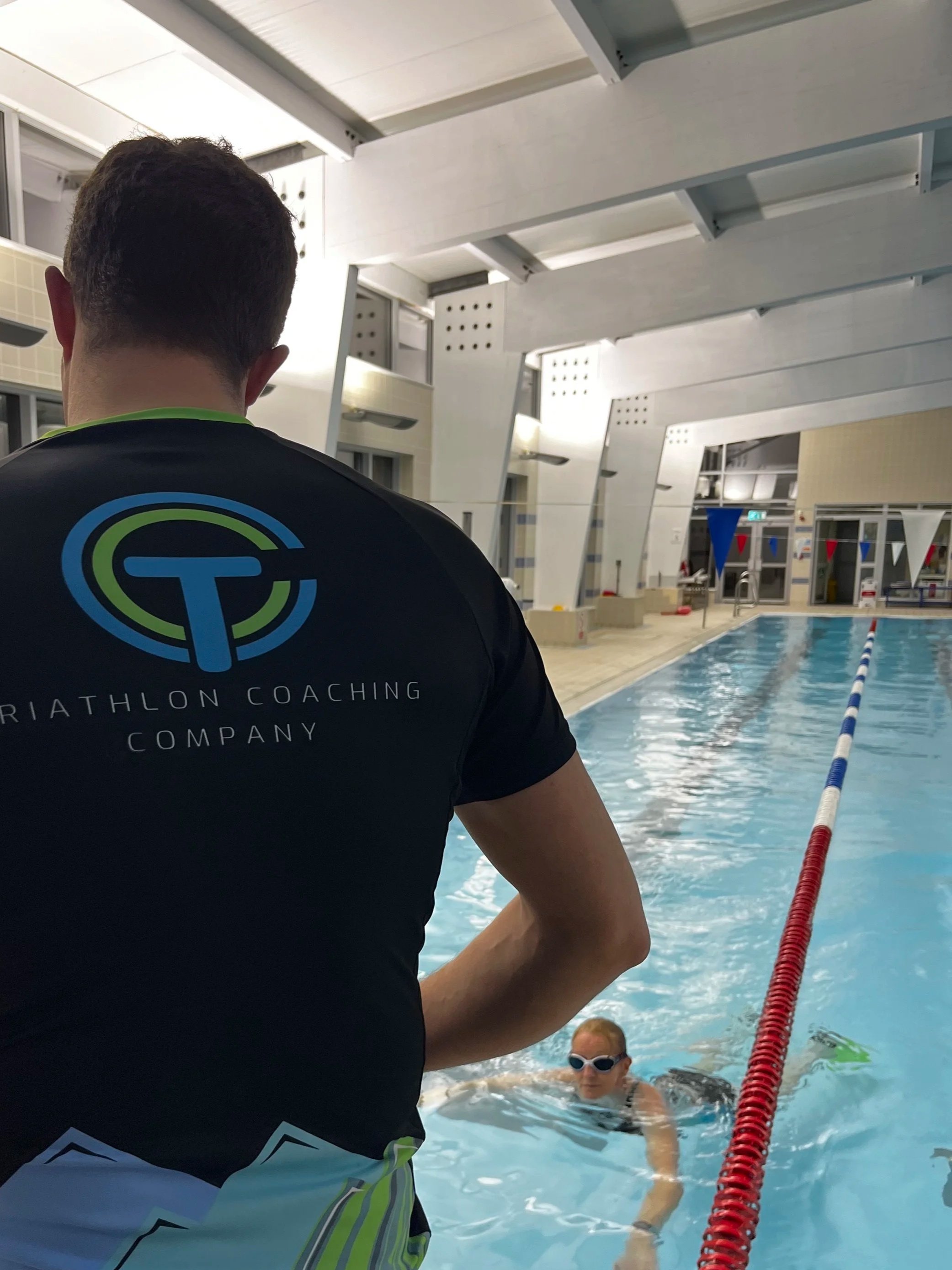Swimmer at an indoor swimming pool with coach observing, pool lane with red and blue lane dividers, swimmer wearing goggles and swimsuit, modern architecture of the indoor pool facility