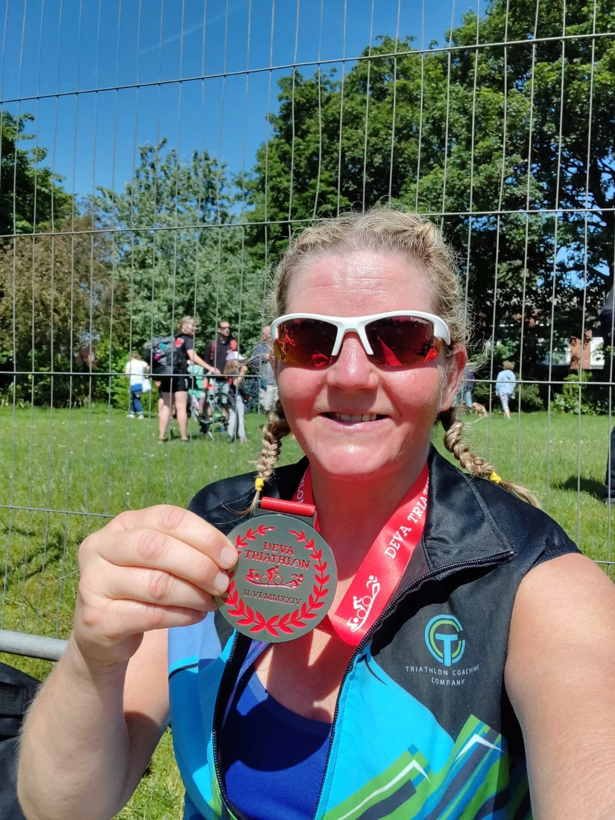 Woman wearing sunglasses holding a silver medal at a triathlon event outdoors with green trees and people in the background.