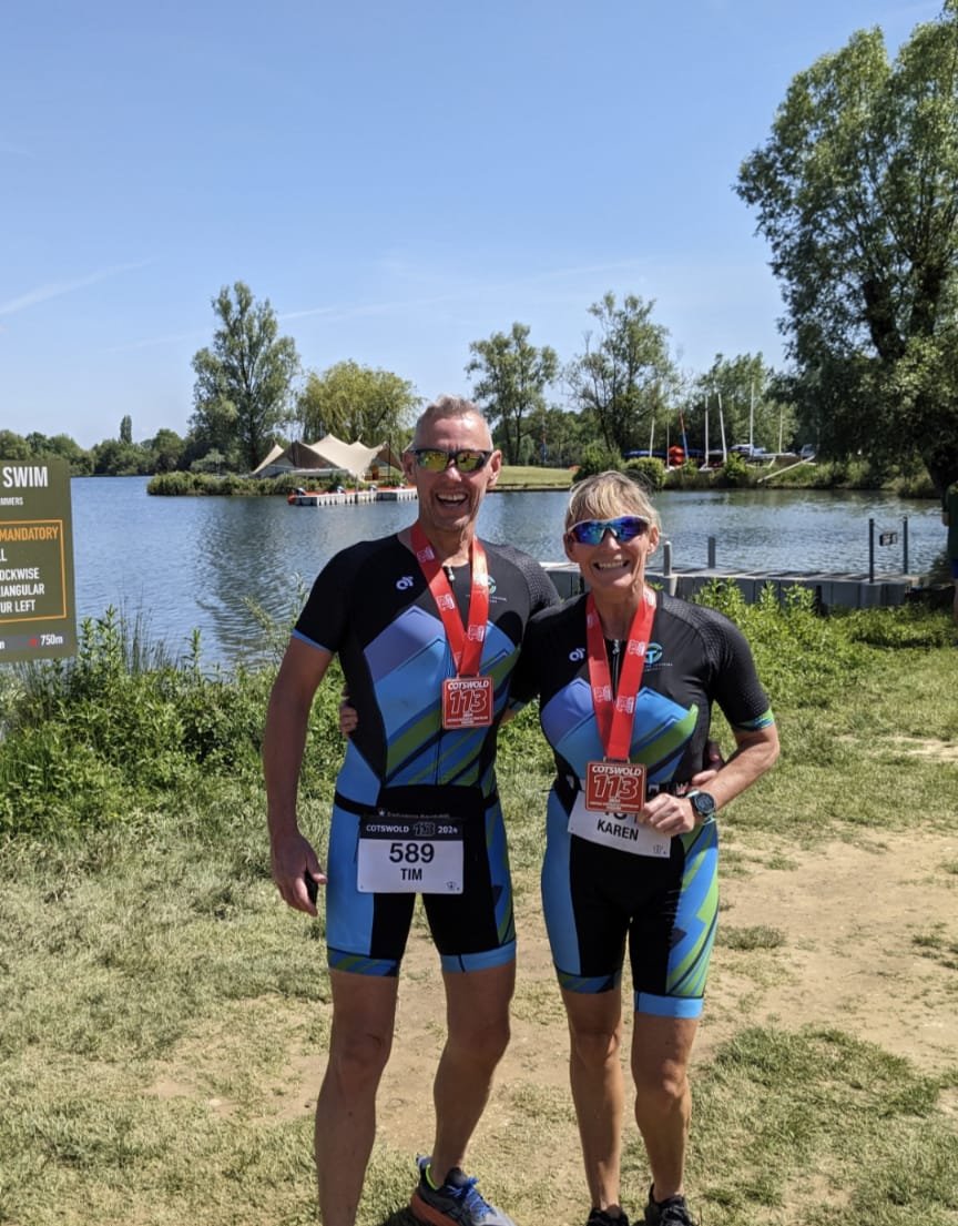 Two smiling runners, a man and a woman, wearing black and blue athletic gear with medals around their necks, standing on grass near a lake with trees and sailboats, after a race.