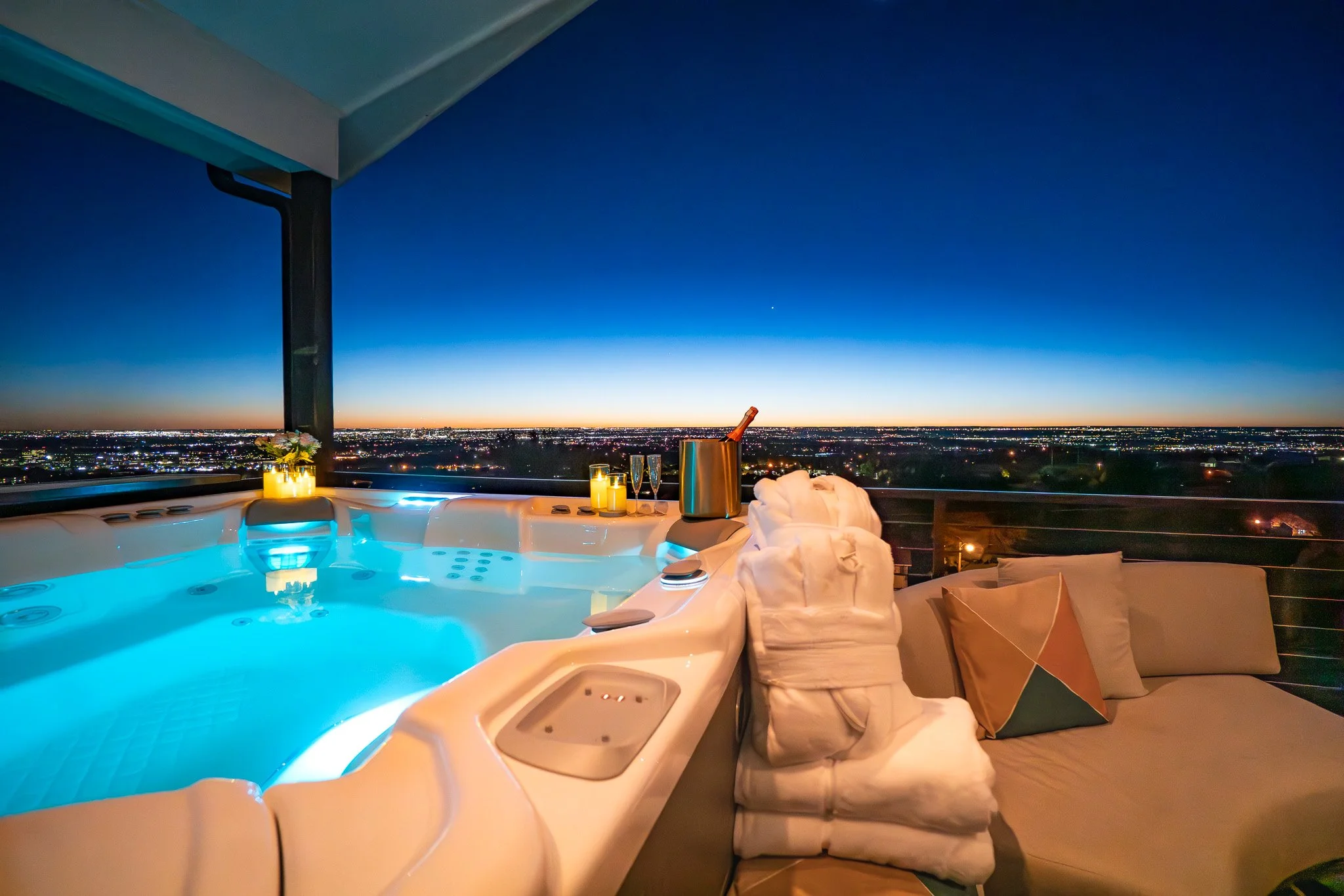 A massaging hot tub with blue water, lit candles, and a view of the Denver cityscape at dusk, with folded towels and a champagne bottle in an ice bucket nearby.