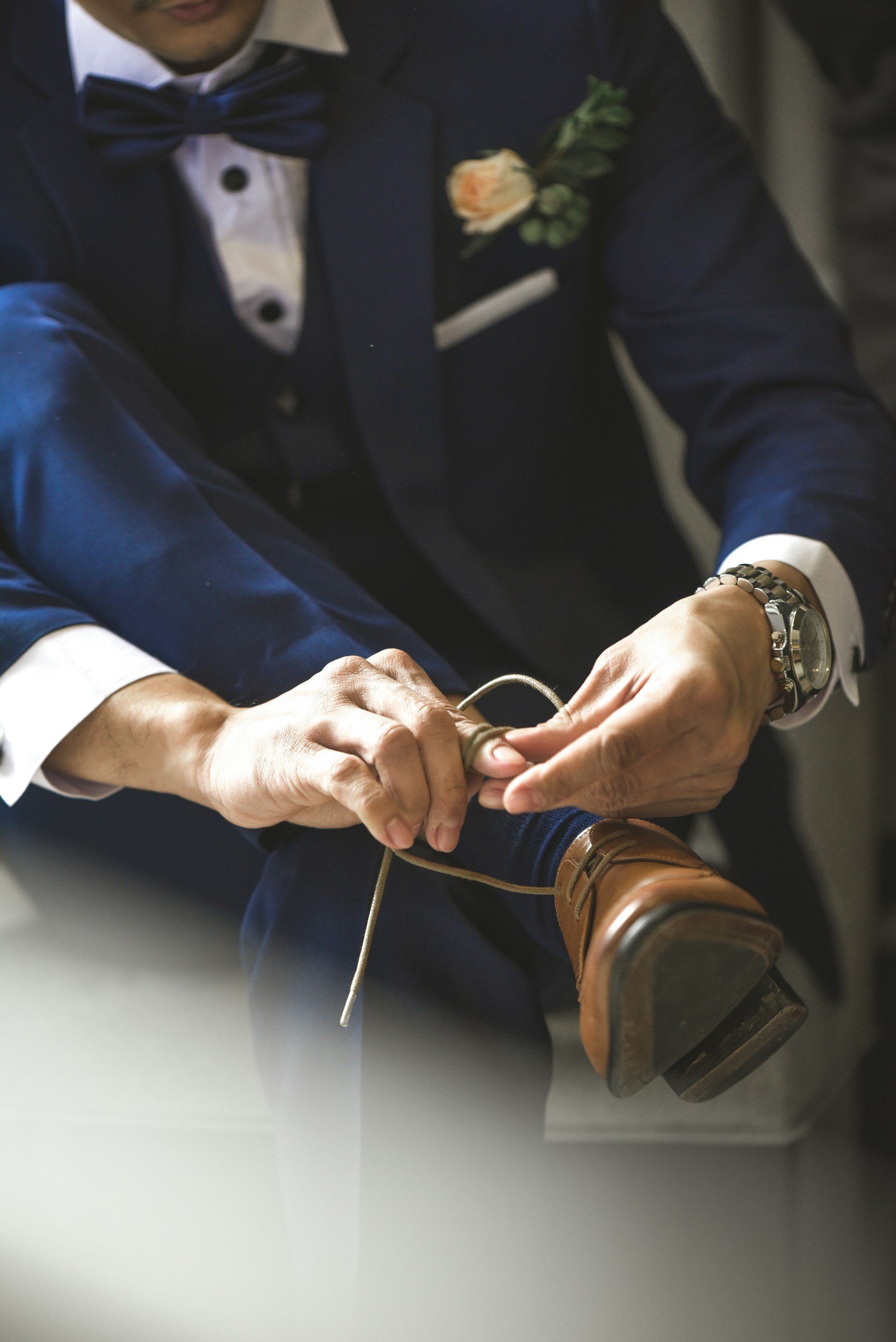 A groom's getting ready photo in a navy blue tuxedo with a white shirt and a bow tie is tying the shoelaces of a brown dress shoe. The man is wearing a wristwatch and has a corsage pinned to his tuxedo in the Honeymoon Suite at Denver Skyline Venue.