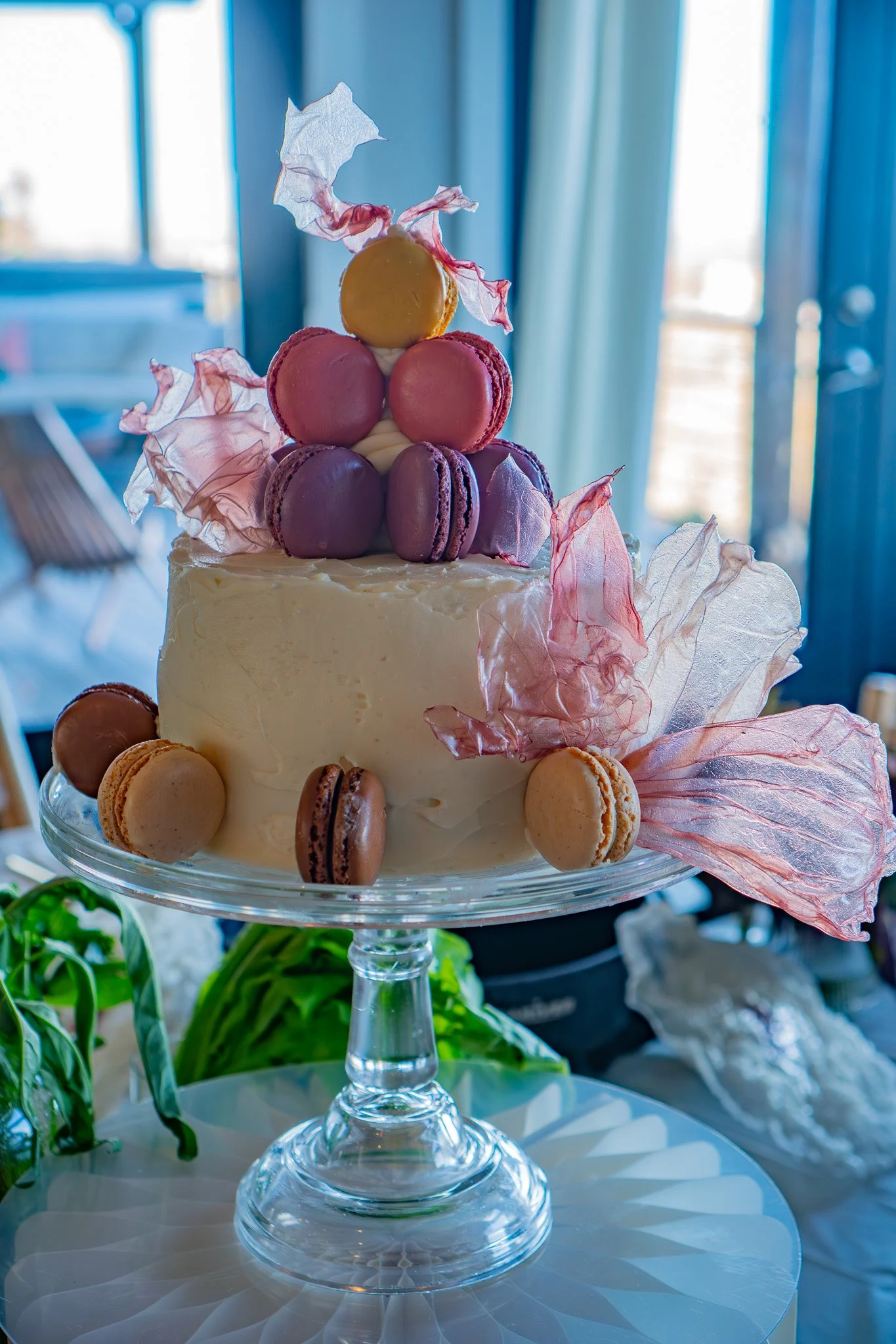 A multi-tiered micro wedding cake decorated with macarons and colorful edible flower petals on a glass cake stand.
