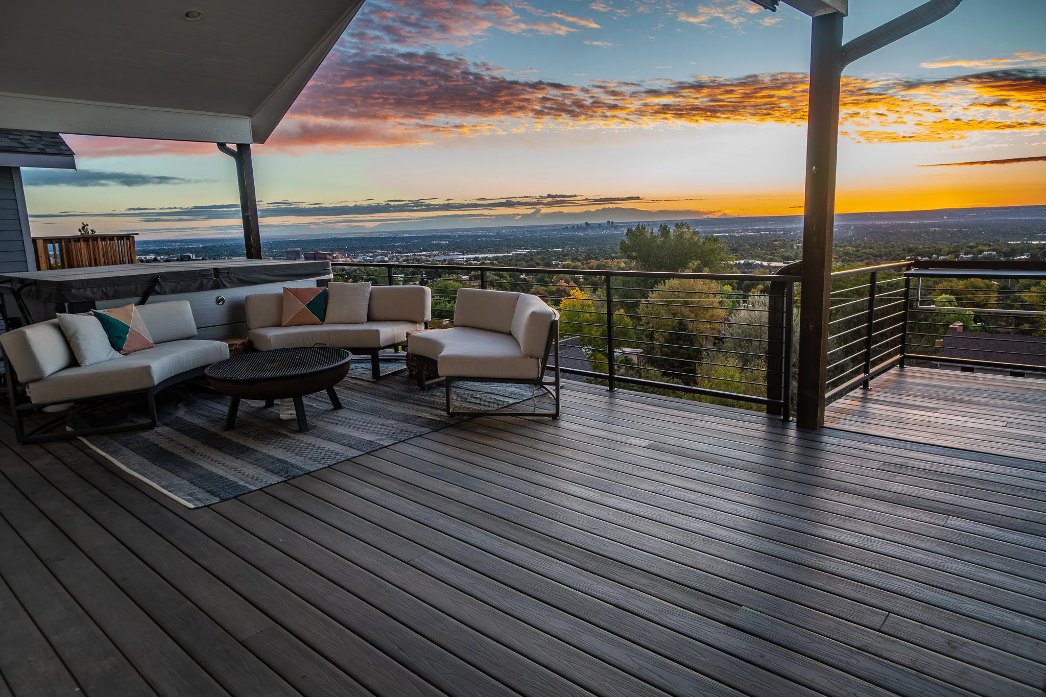 A spacious wooden deck with outdoor furniture, including a white sofa, armchair, and coffee table, overlooking a scenic Denver city view during sunset with colorful clouds in the sky.