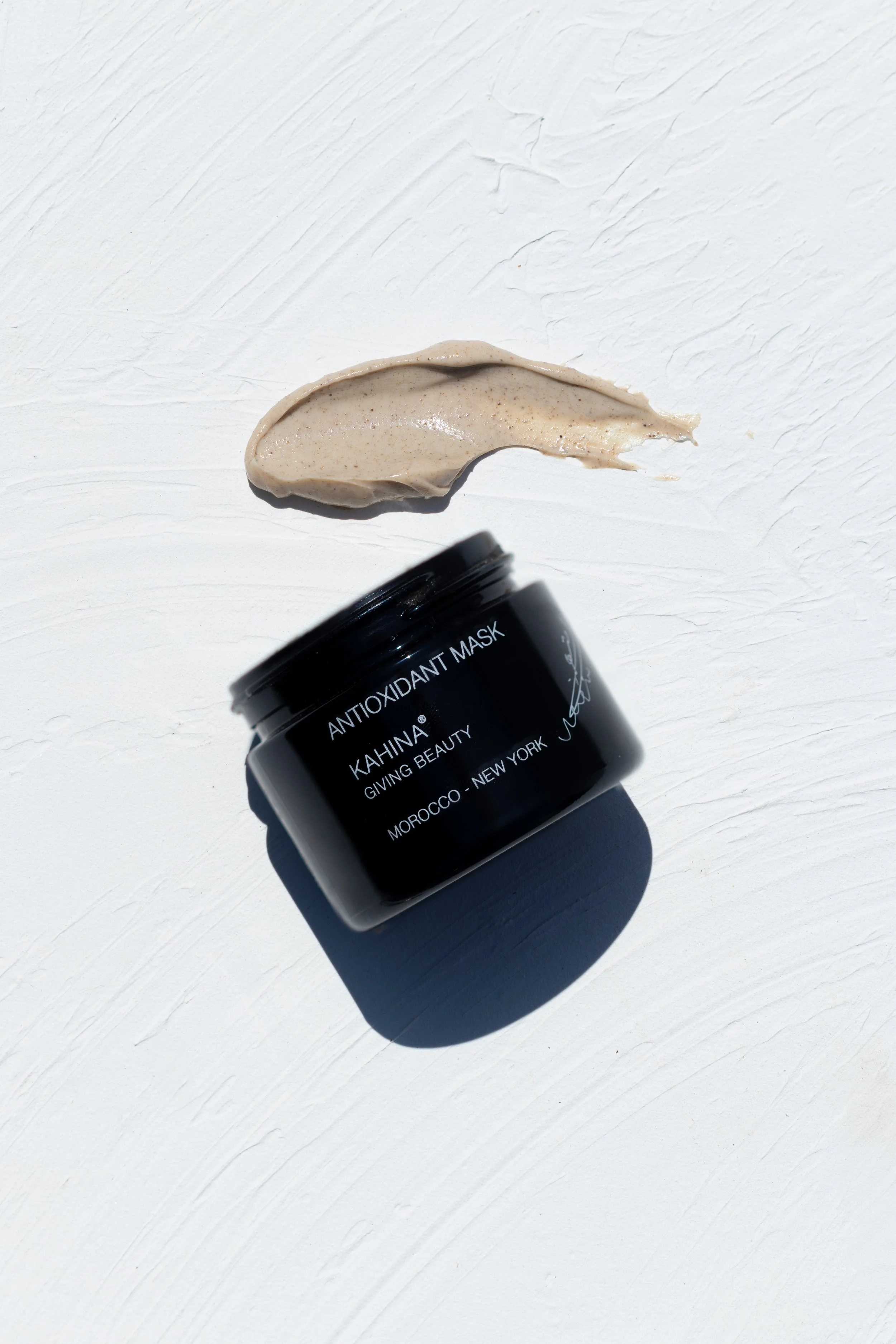 Open black jar of Kahina Antioxidant Mask with a dollop of product on white textured surface.