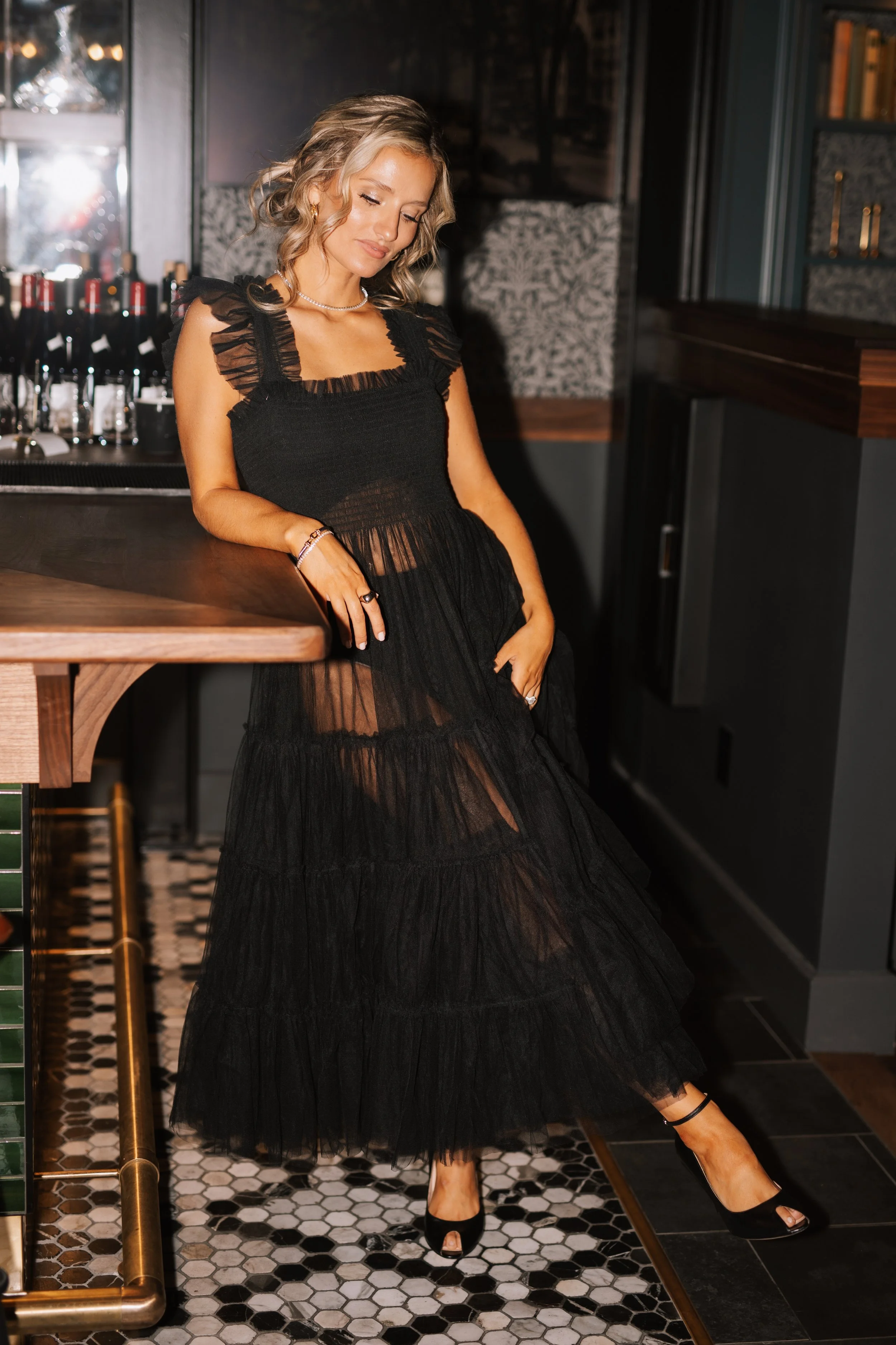 A woman in a sheer black dress with ruffles on the shoulders, wearing black high heels, standing in a dimly lit bar or restaurant interior.