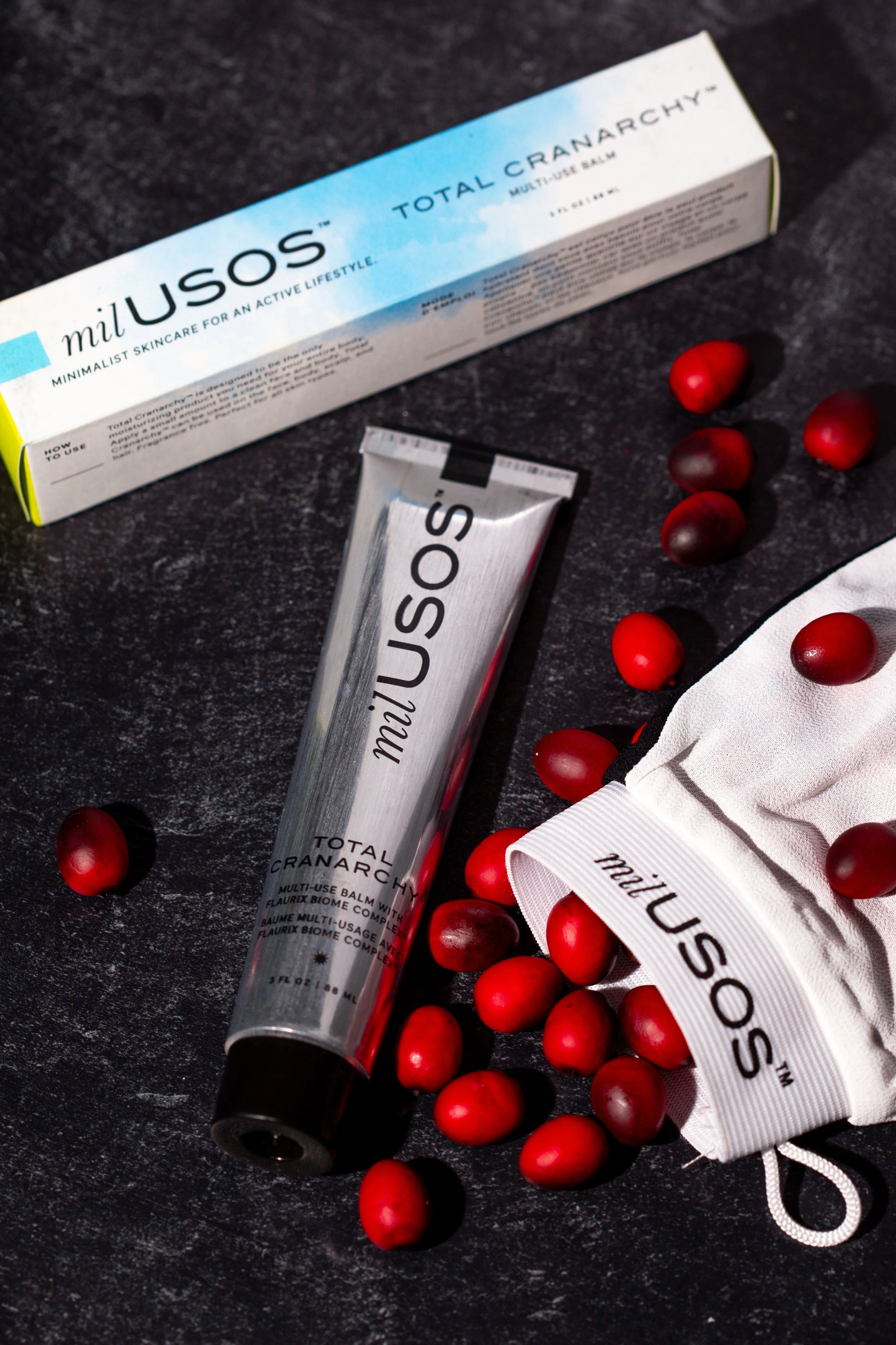 A tube of milUSOS Total Cranarchy multi-use balm, a white cloth bag with the milUSOS logo, scattered red berries, and the balm box on a dark textured surface.