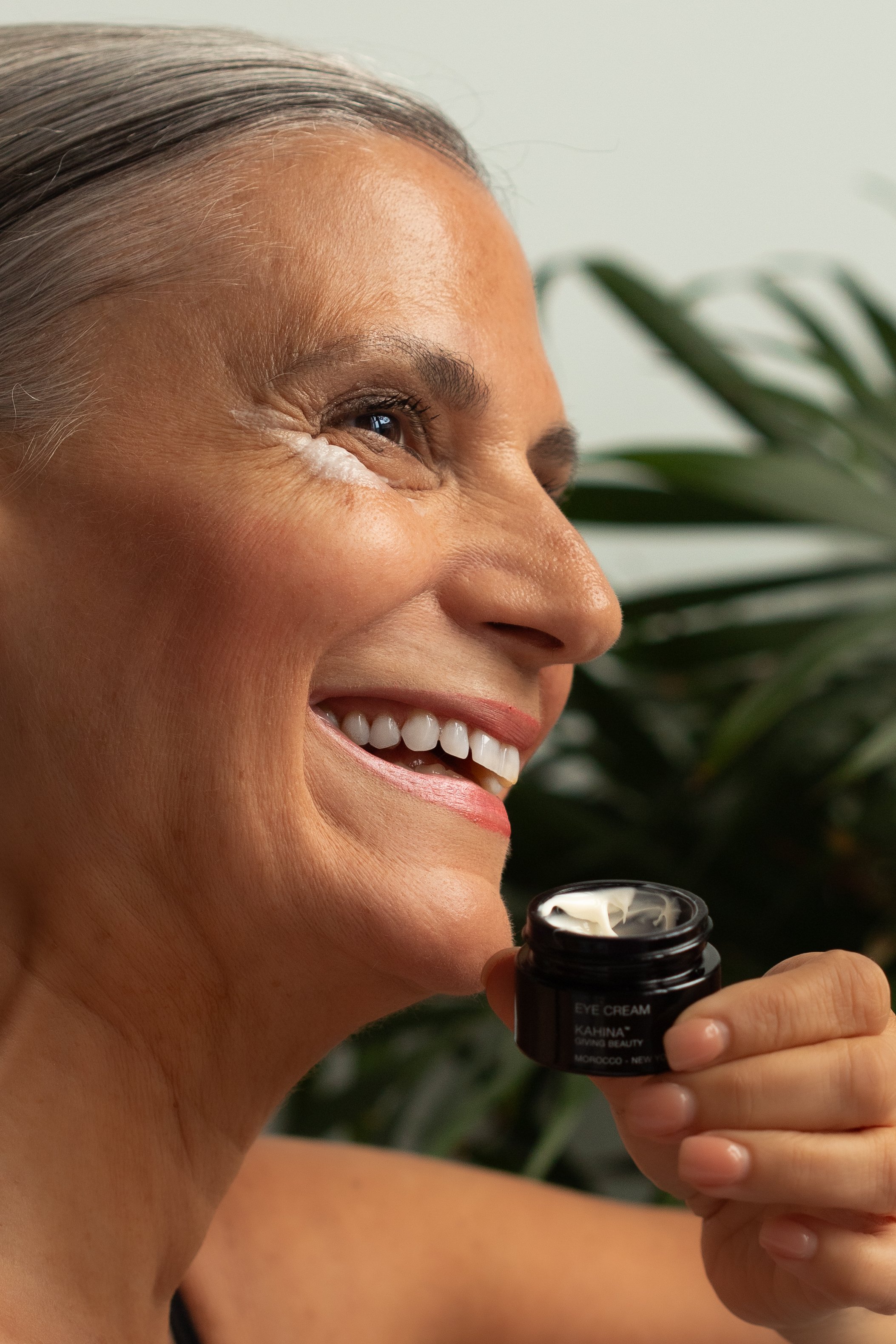 A woman smiling while holding a small black jar of eye cream near her eye, with greenery in the background.