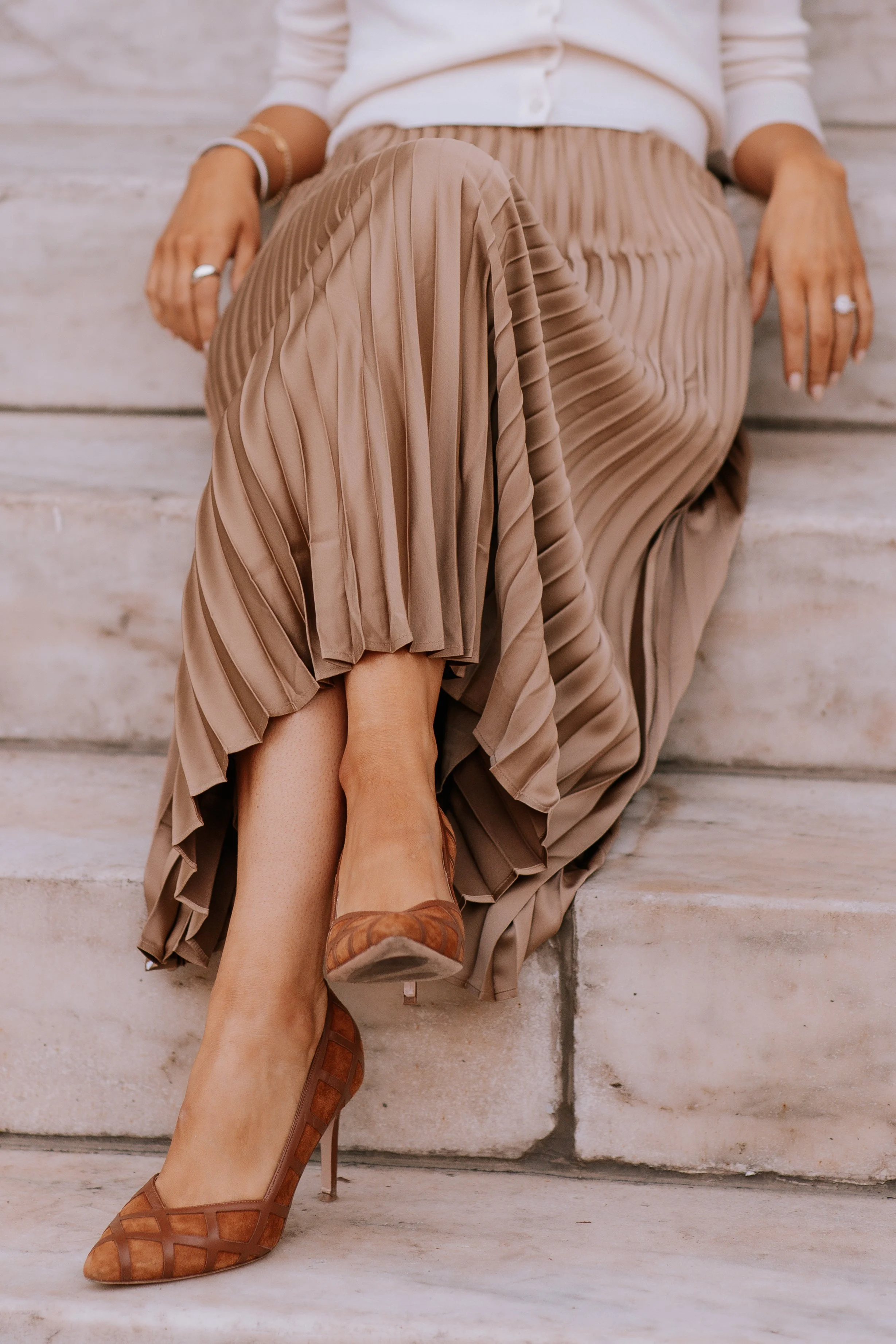A woman wearing a white top, beige pleated skirt, and high heels, sitting on stone steps with her legs crossed.