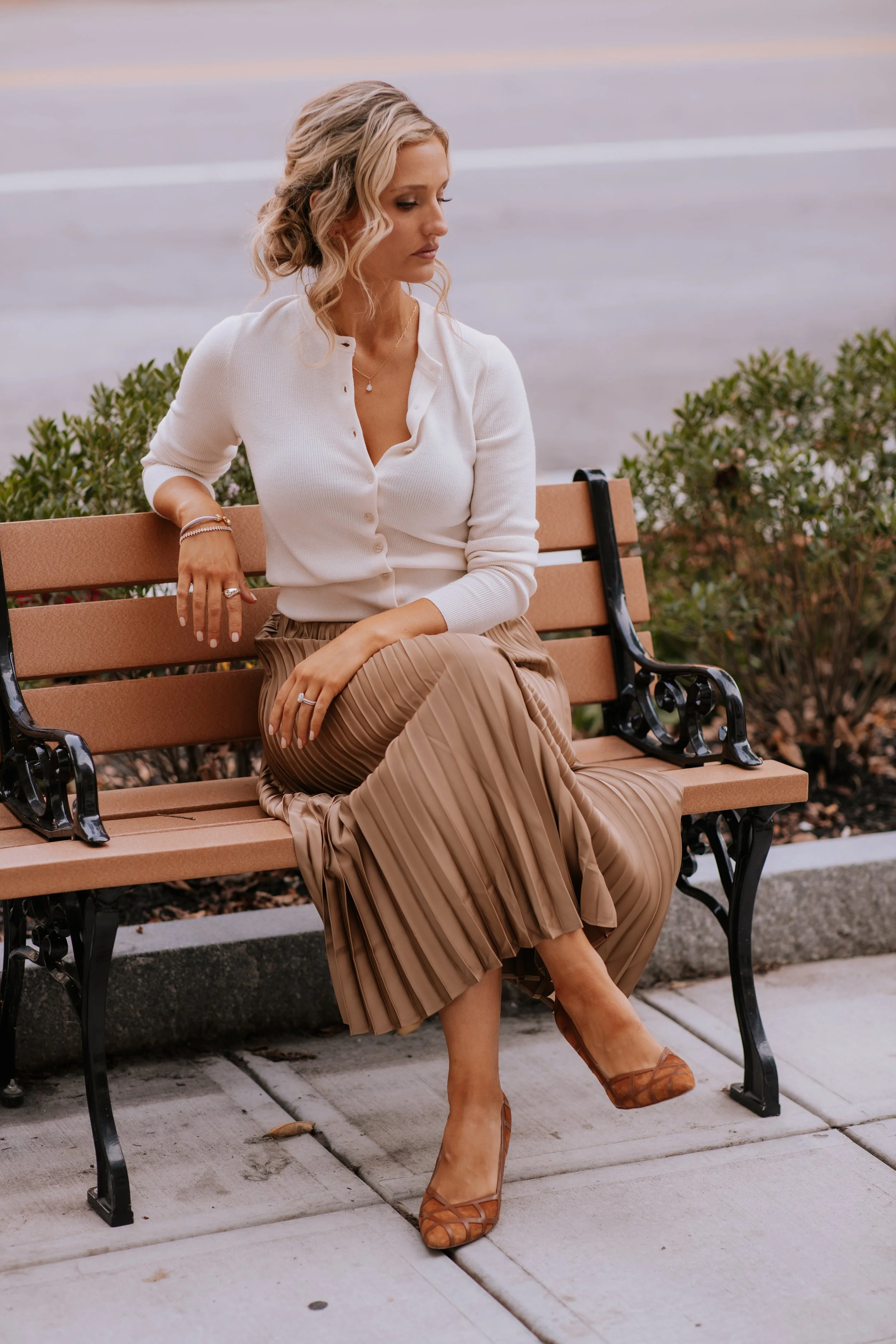 A woman with blonde hair styled in loose waves, wearing a white button-up cardigan, a pleated tan skirt, and brown heels, sits on a park bench looking to the side.