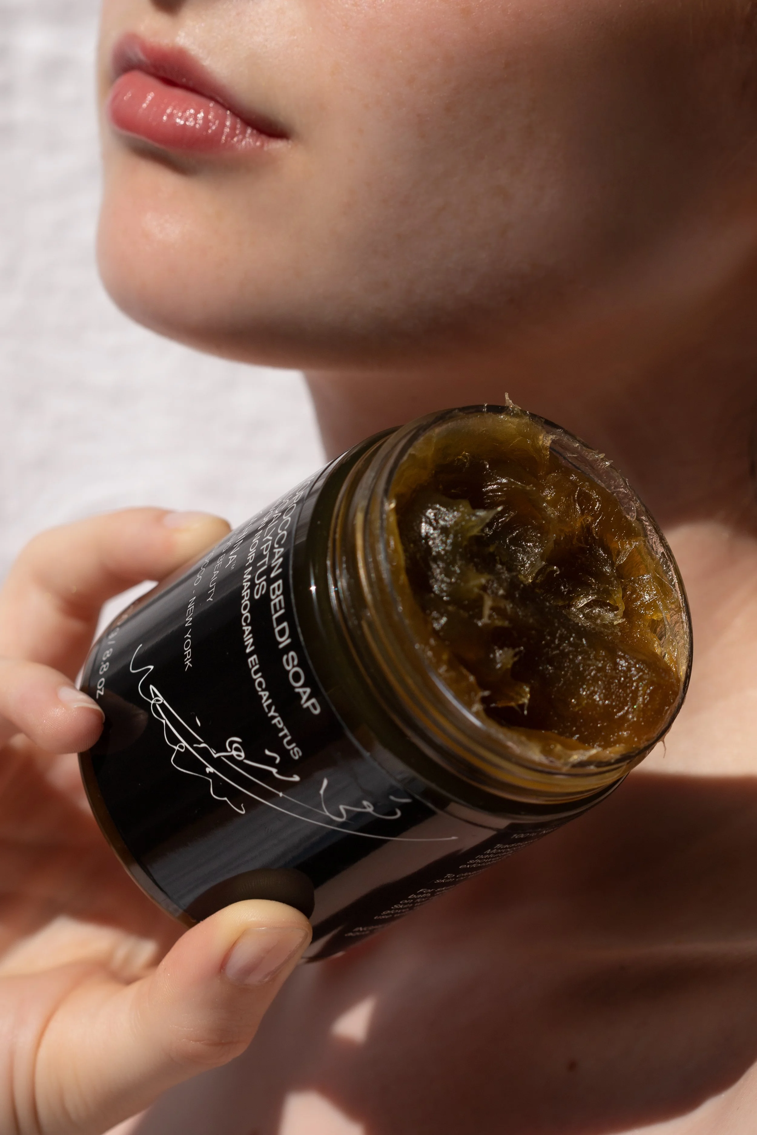 Close-up of a woman's face near her lips, holding an open jar of skincare or hair product, partially visible. The jar contains a gel-like substance with a gold-brown tint.