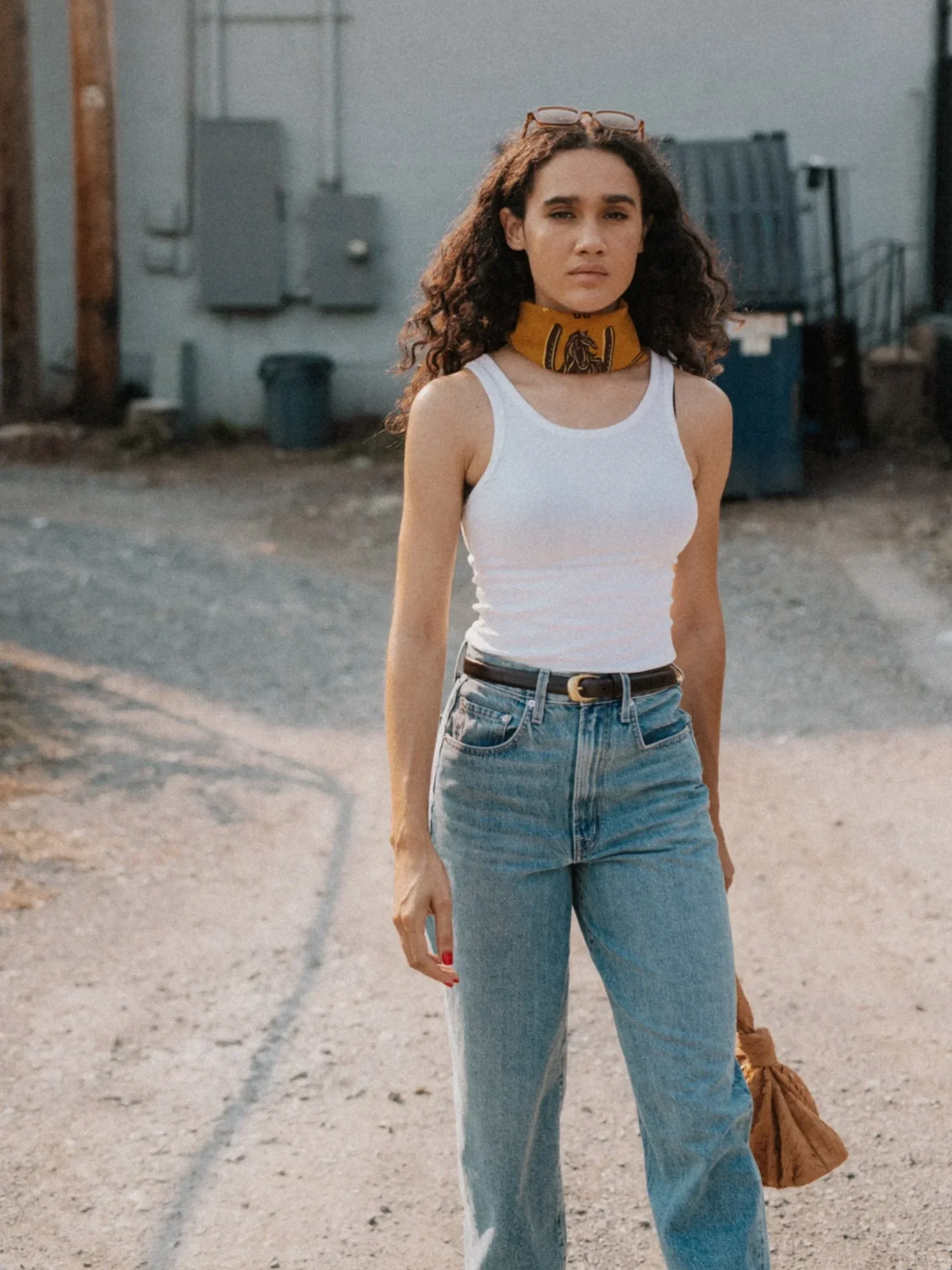 A young woman with curly hair wearing a white tank top, high-waisted blue jeans, and a black belt. She has sunglasses perched on her head and a yellow bandana with a horse design around her neck. She stands outside on a gravel surface, with a serious expression.