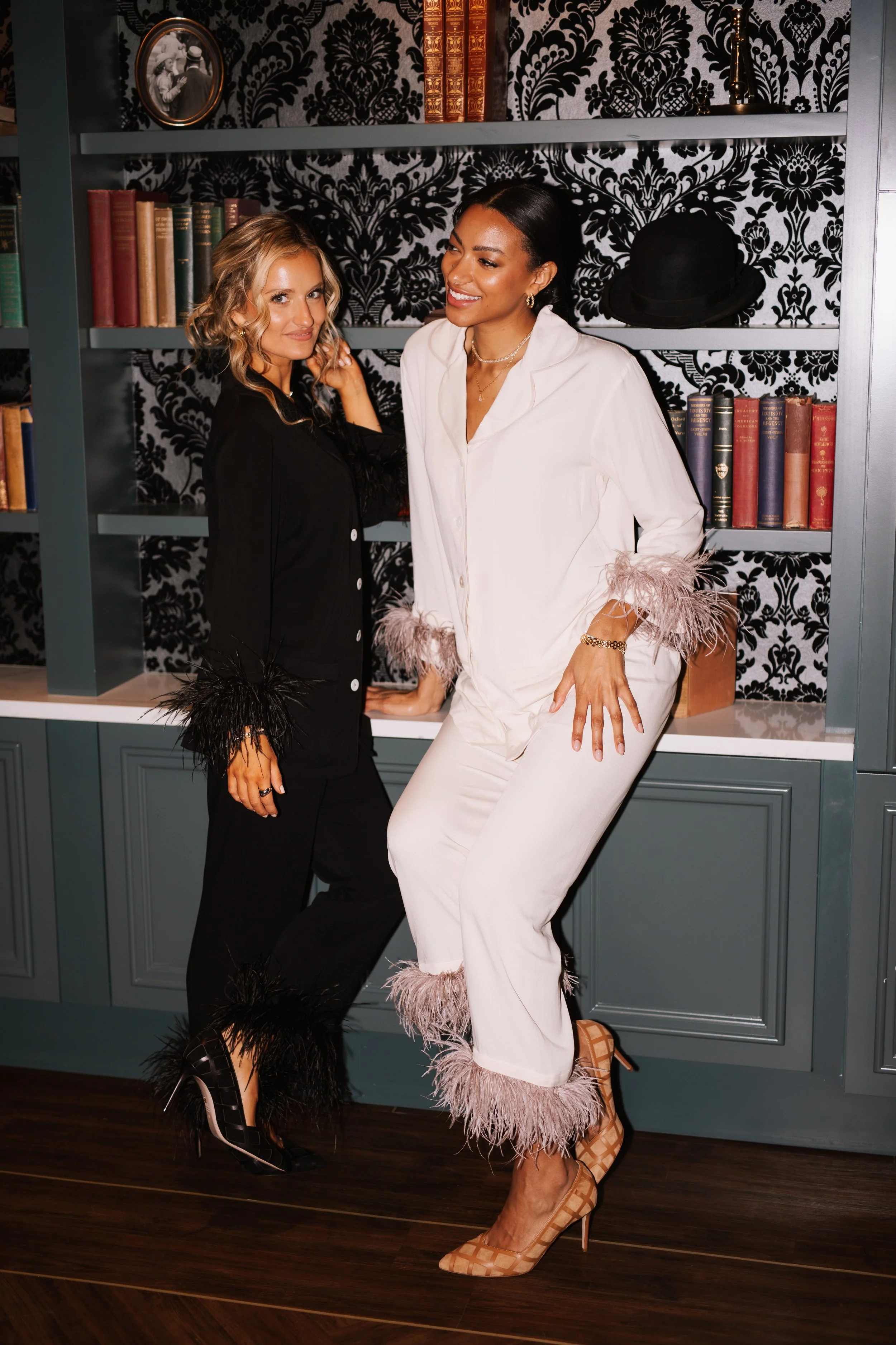 Two women posing in front of a bookshelf with vintage books and a framed photo. One woman is dressed in black with feathered cuffs, and the other in white with feathered cuffs and pants, wearing high heels.