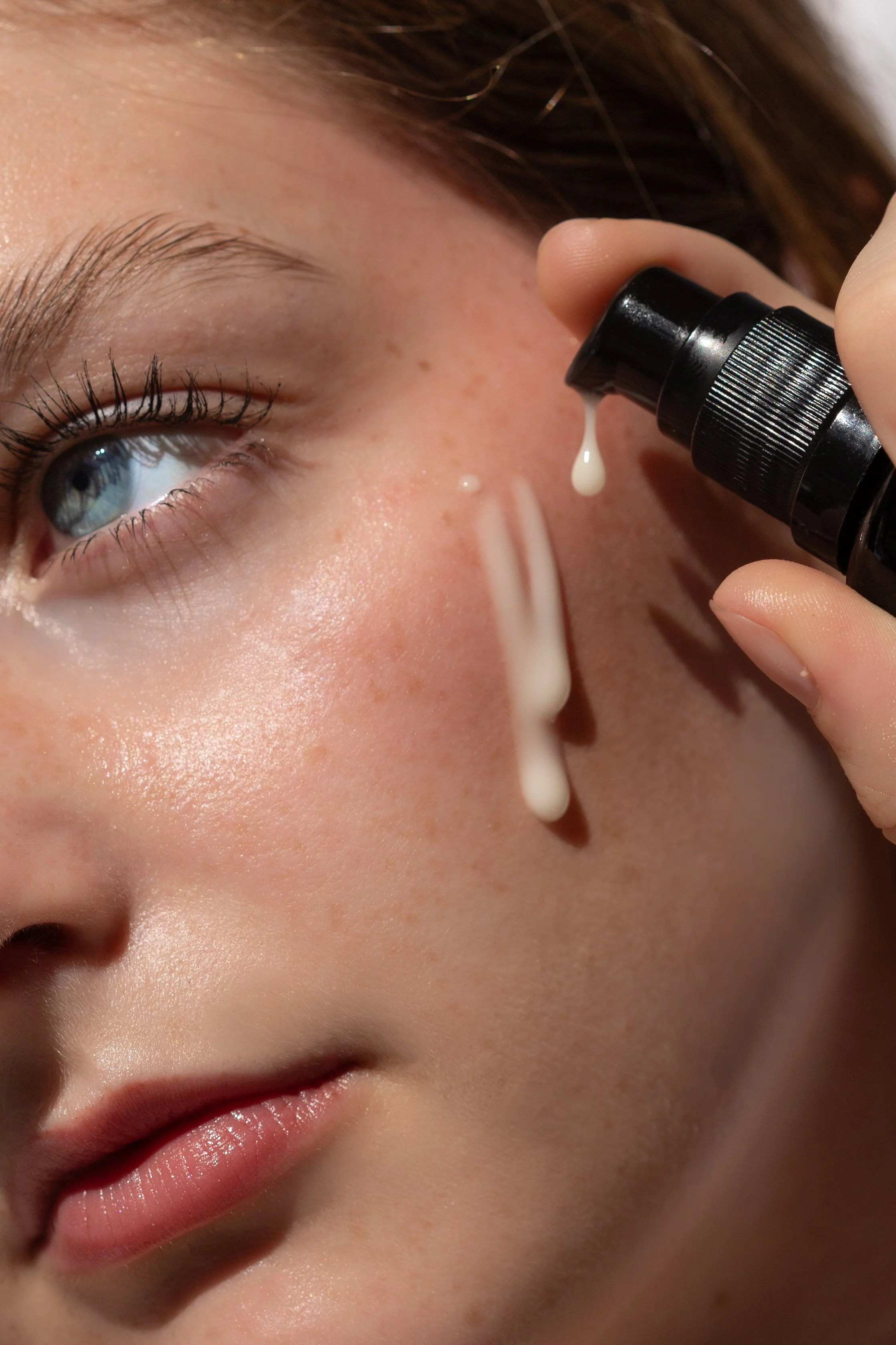 Close-up of a woman with fair skin, blue eyes, and light makeup, applying white cream or lotion under her eye from a black pump bottle.