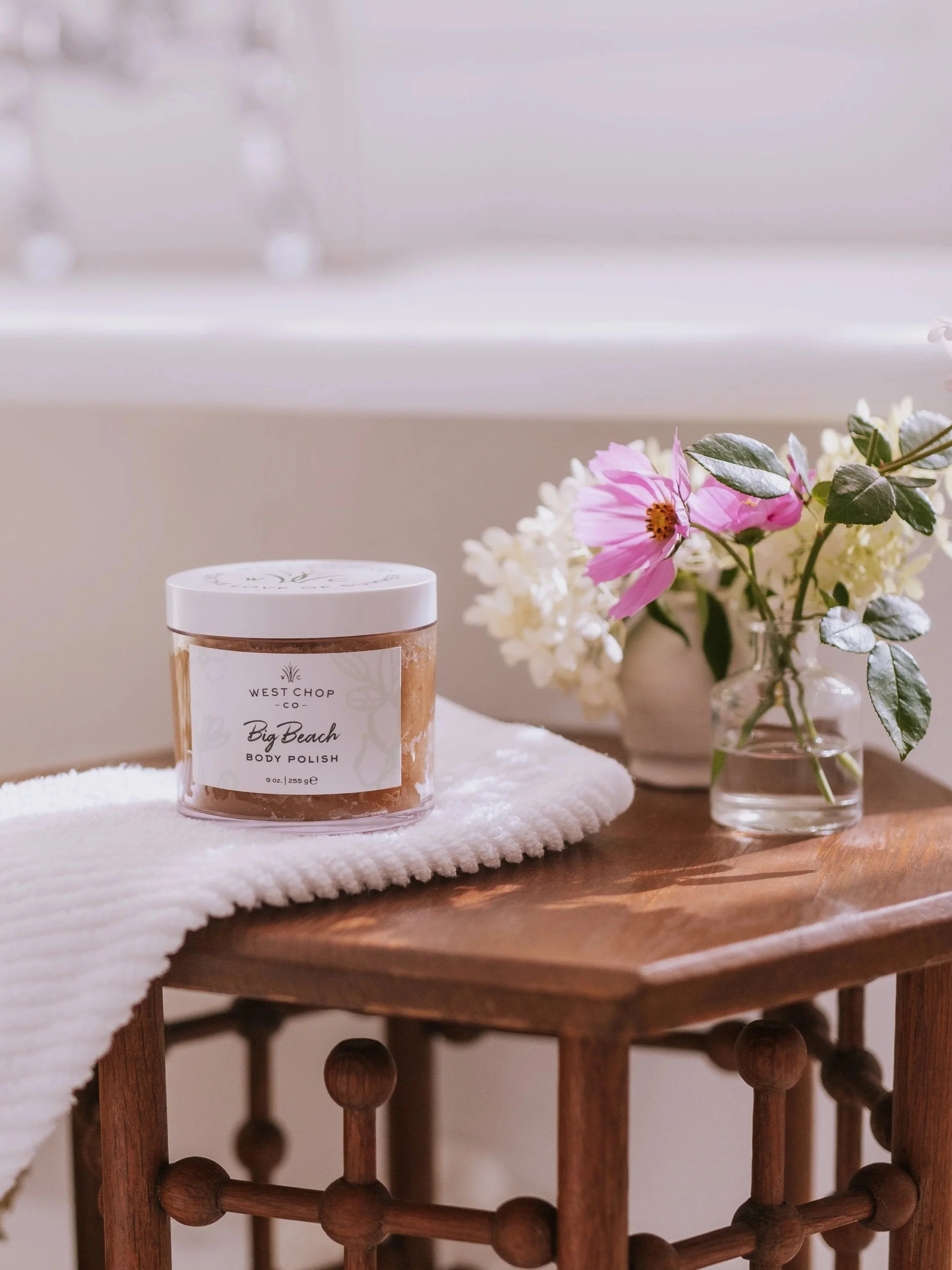 A jar of West Chop Co Big Beach body polish on a white towel on a wooden table, next to a small vase with pink and white flowers, near a window.