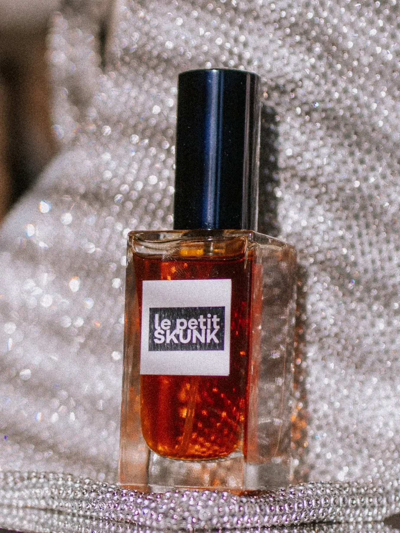 A perfume bottle labeled 'le petite SKUNK' placed on a sparkling, silver, beaded fabric background.