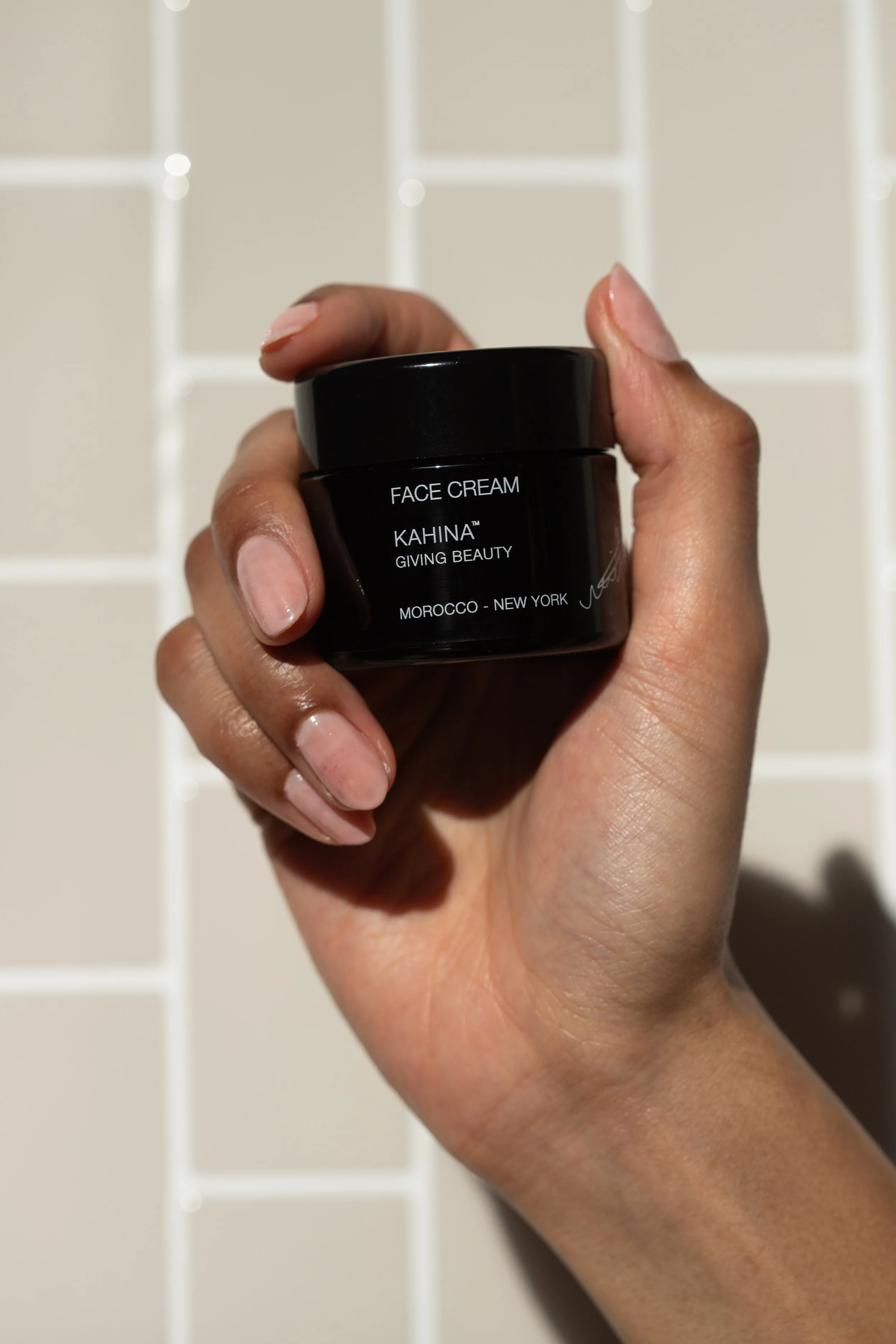 A hand holding a small black jar of face cream with white text, against a tiled bathroom wall background.