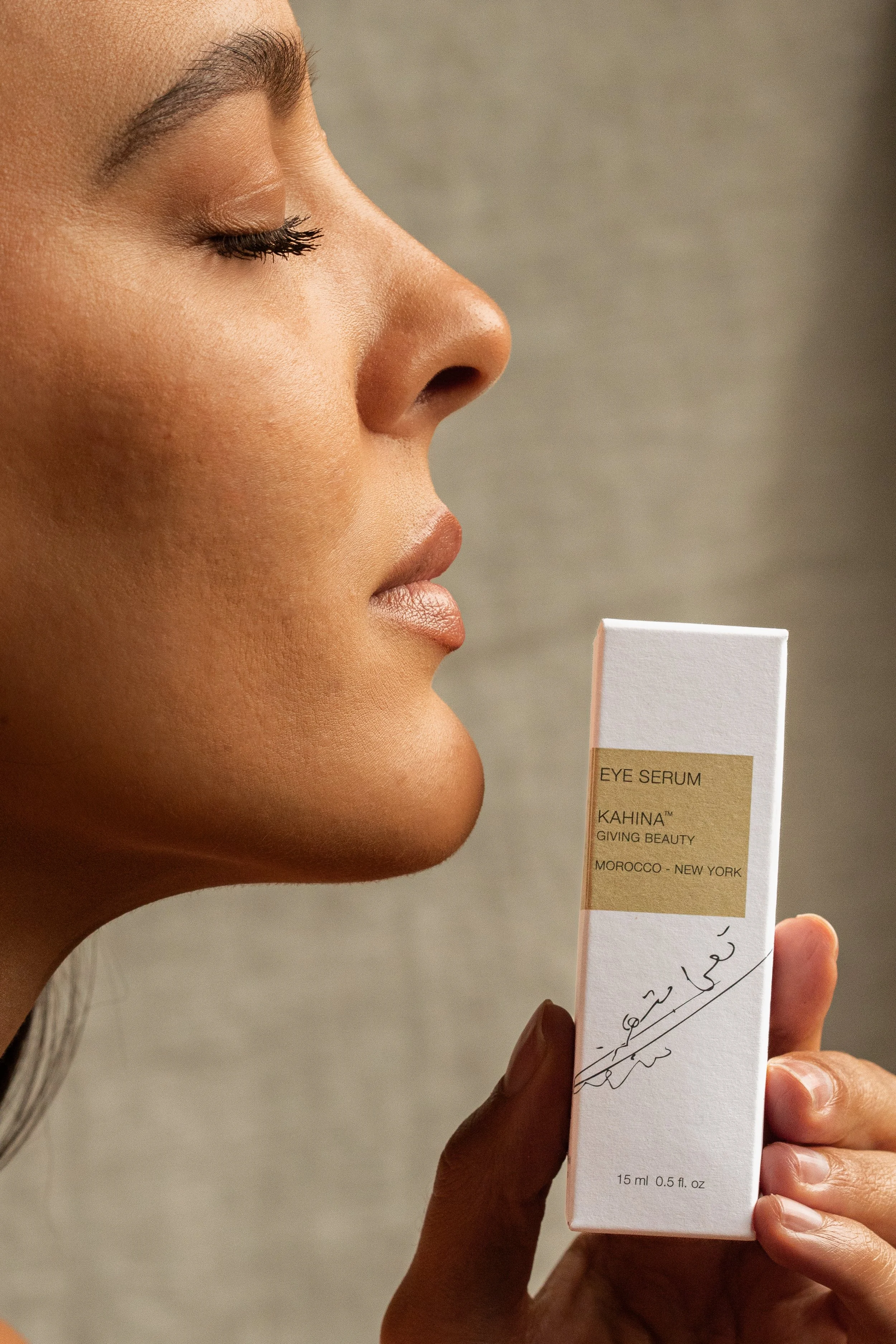 A woman holding a white box of eye serum with Arabic writing and English text, with her face turned sideways and eyes closed.