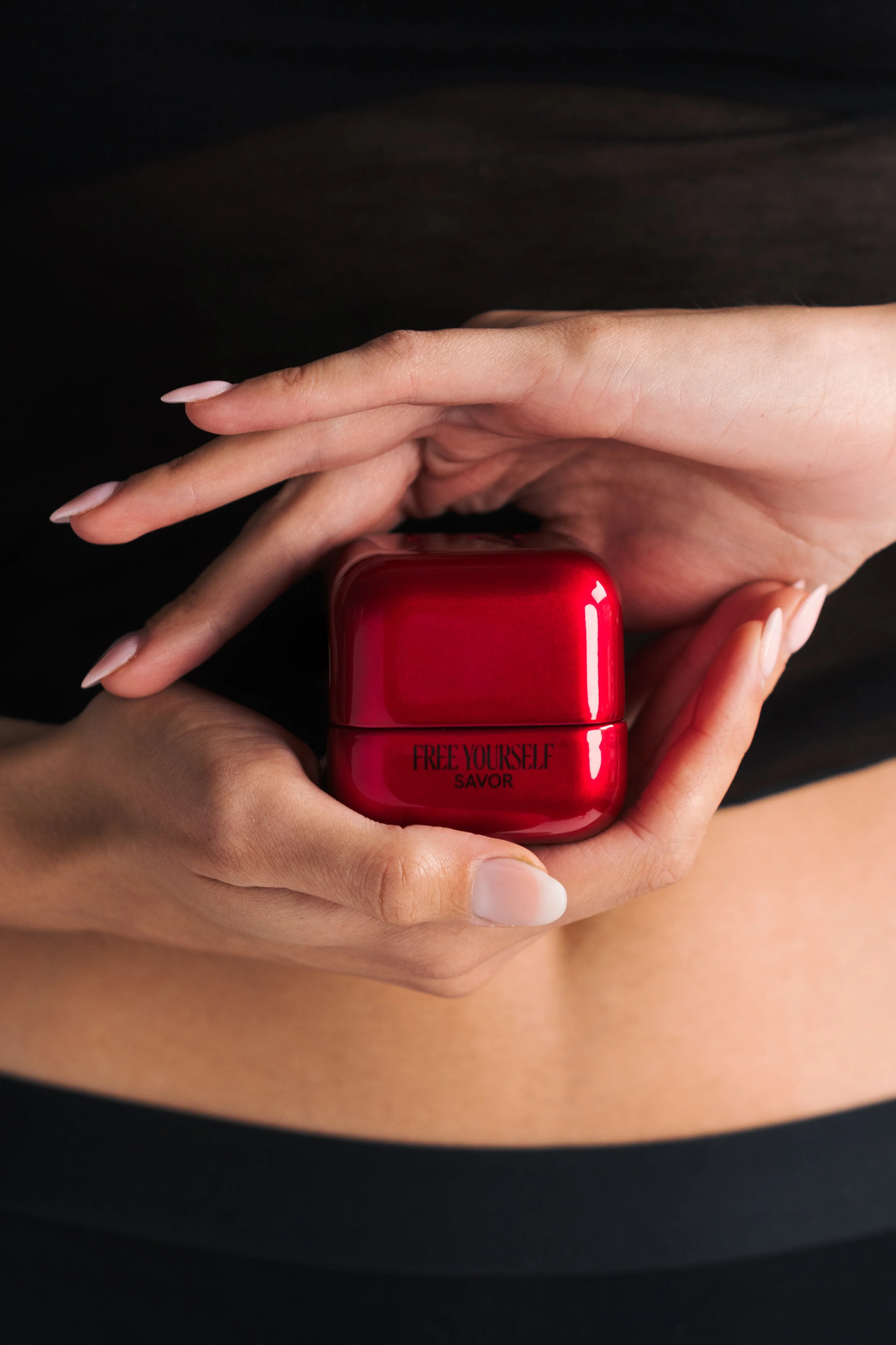 Close-up of hands holding a red cosmetic container with the label 'FREE YOURSELF SAVOR' against a dark background.
