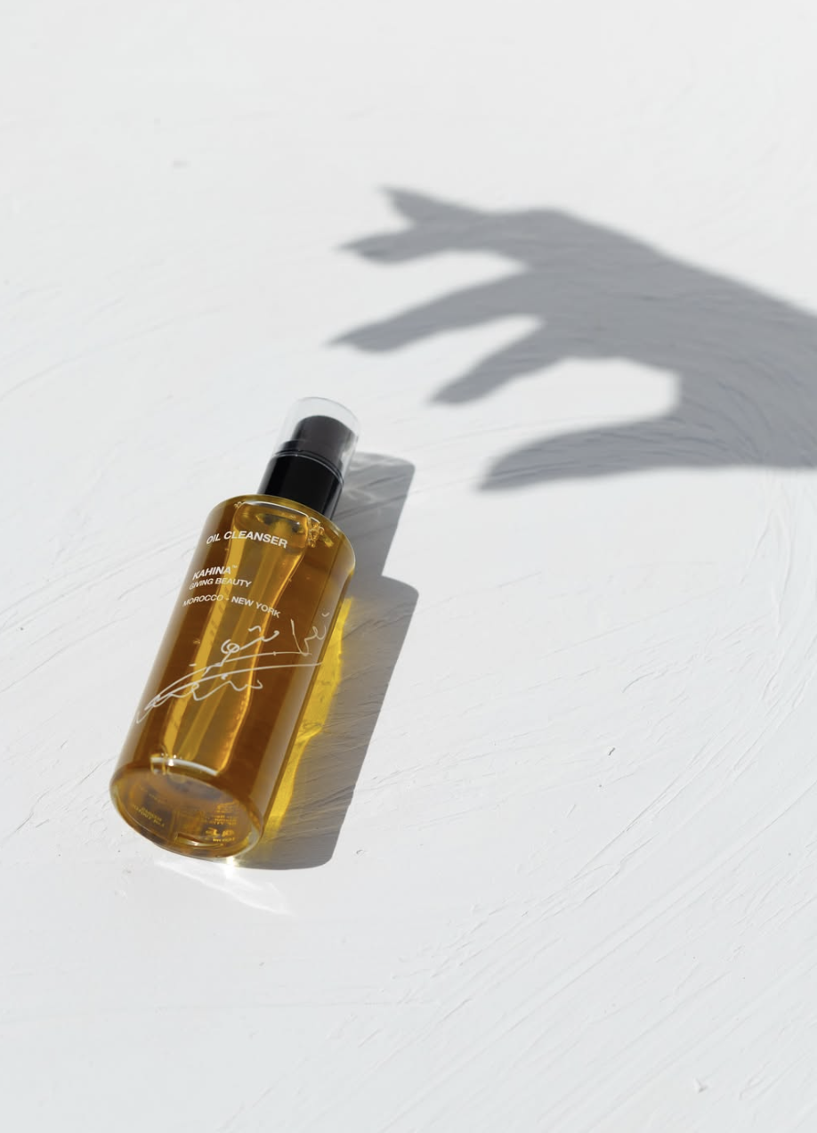 A yellow oil cleanser bottle with a black pump spray top on a white surface, casting a shadow resembling a hand.