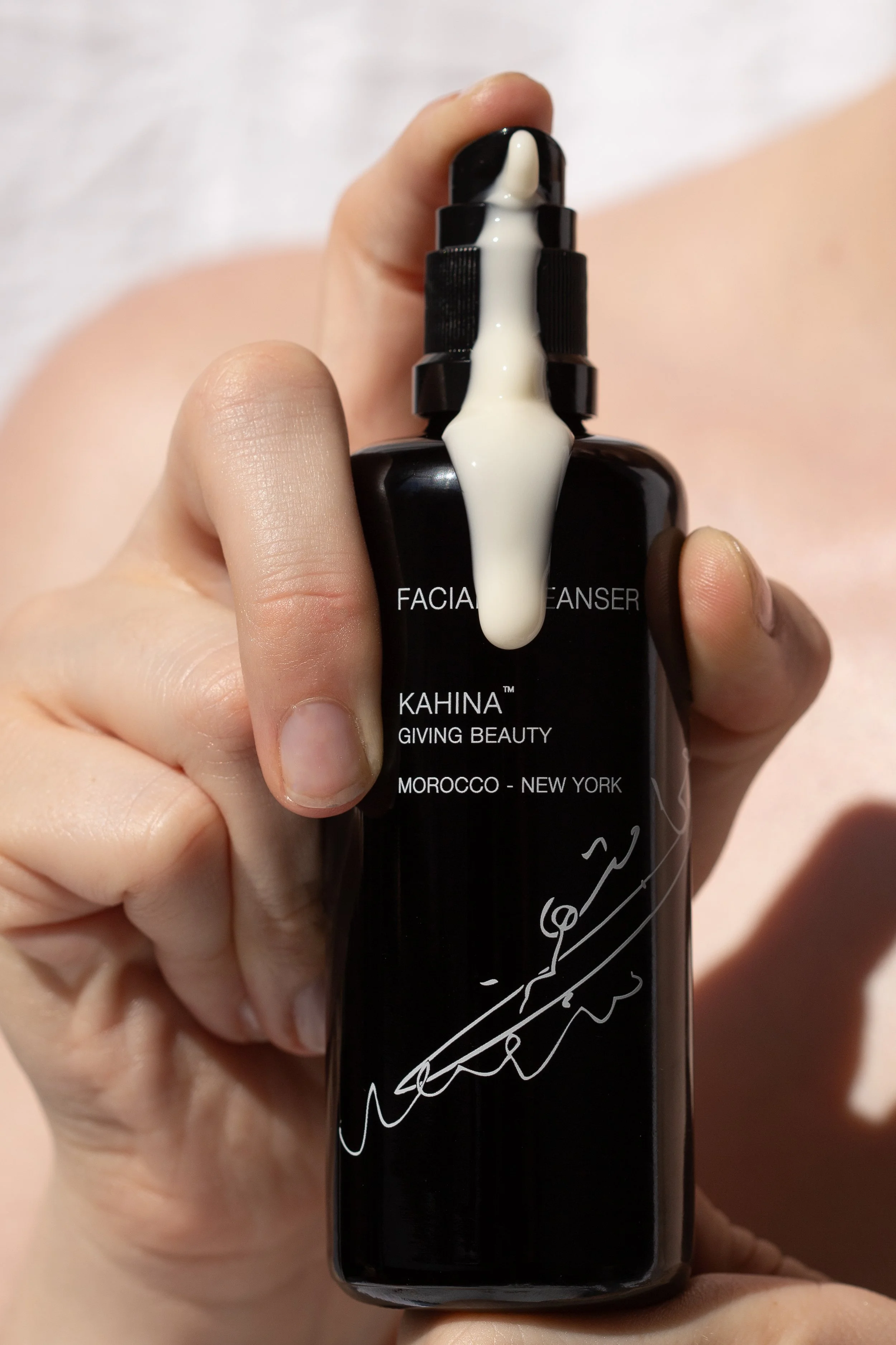 A person's hand holding a black facial cleanser bottle with white text and signature. White product is dispensing from the pump onto the finger.