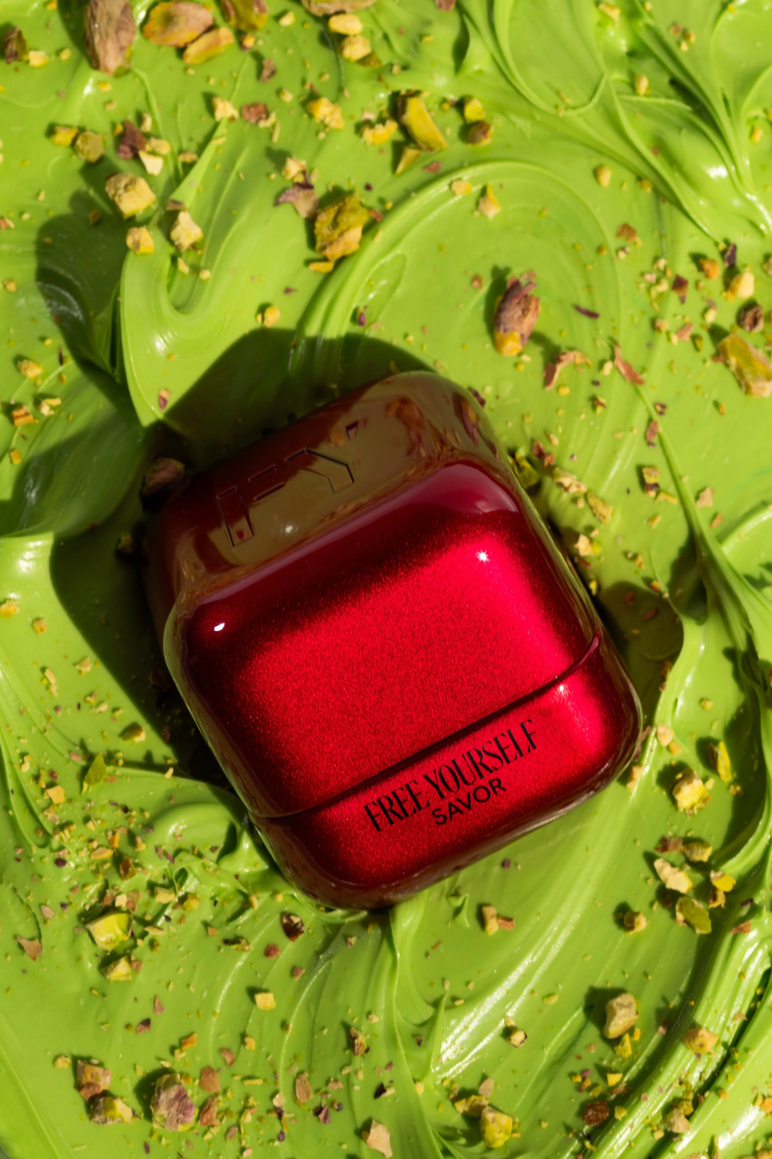 A red and gold perfume bottle labeled 'FREE YOURSELF' resting on large green leaves and crushed pistachios.