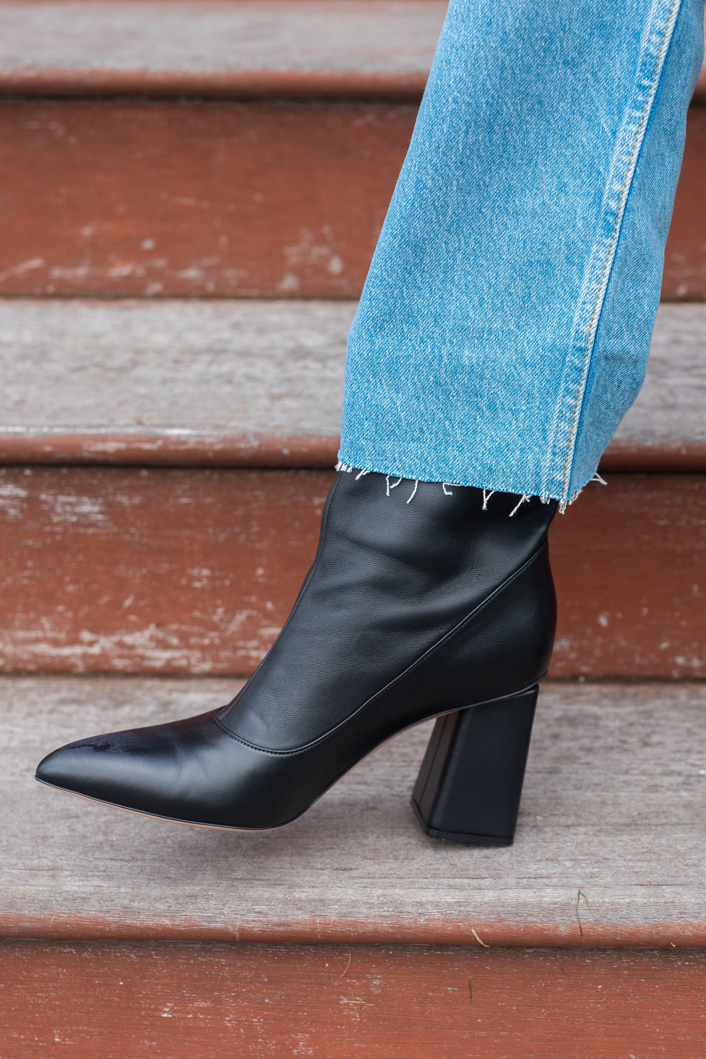 Close-up of a person's foot wearing a black pointed-toe high heel boot with a chunky heel, paired with cuffed blue jeans, standing on wooden steps.