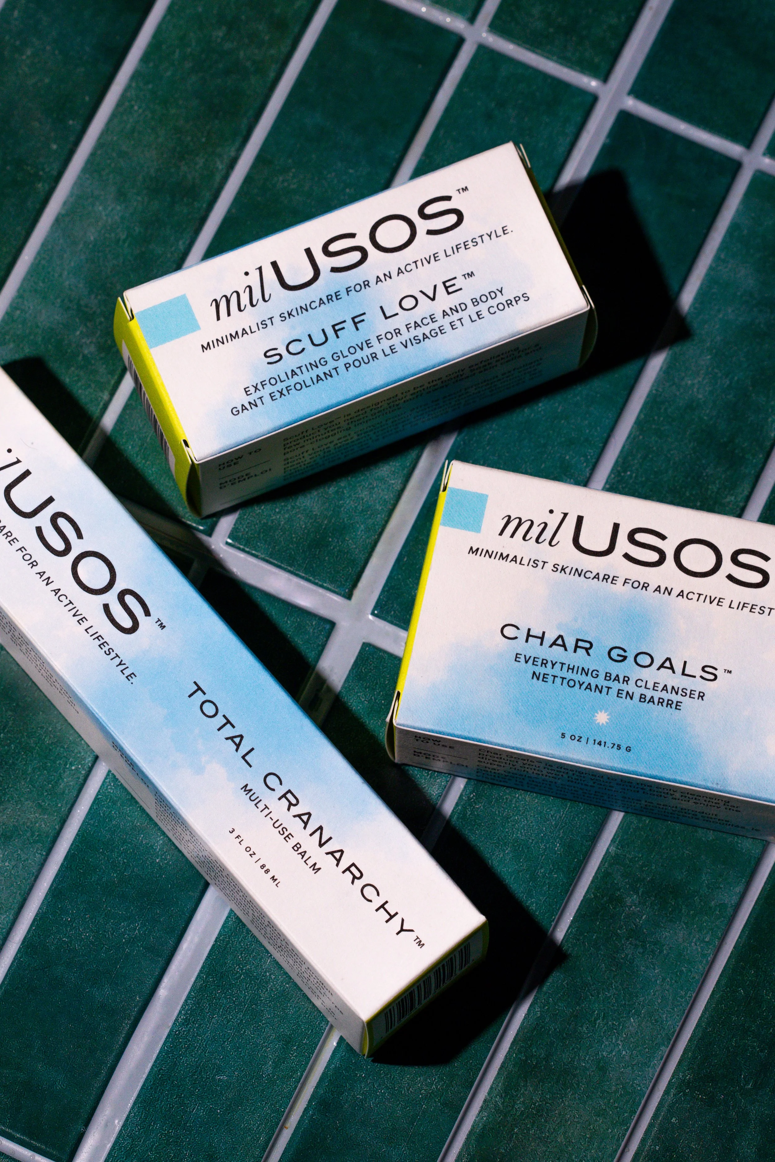 Three skincare product packages on a green tile surface, including Milusos Scuff Love exfoliating face and body glove, Char Goals bar cleanser, and Total Cranarchy multi-use balm.