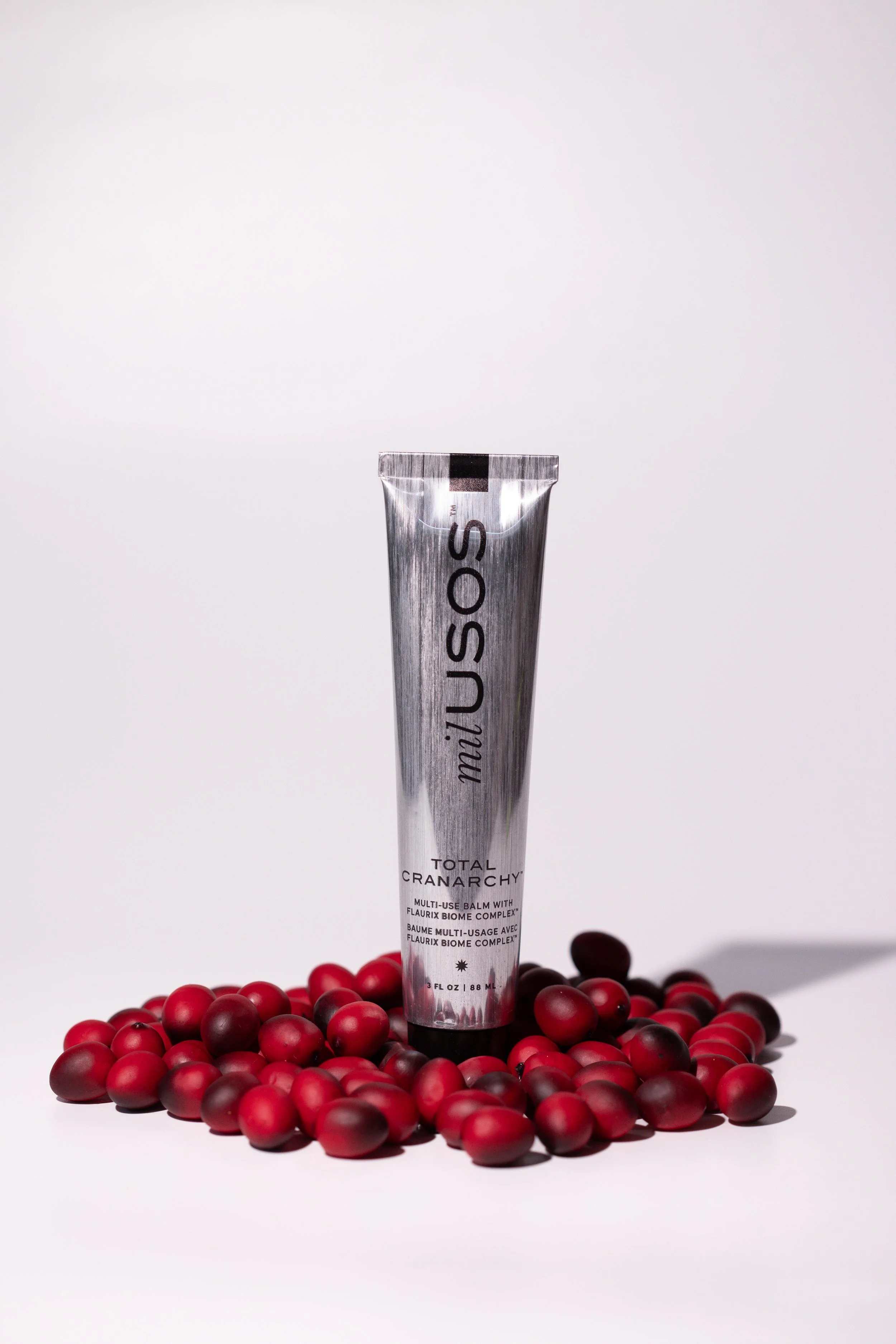 A silver tube labeled "meûUSO" standing upright surrounded by red and black berries on a white background.
